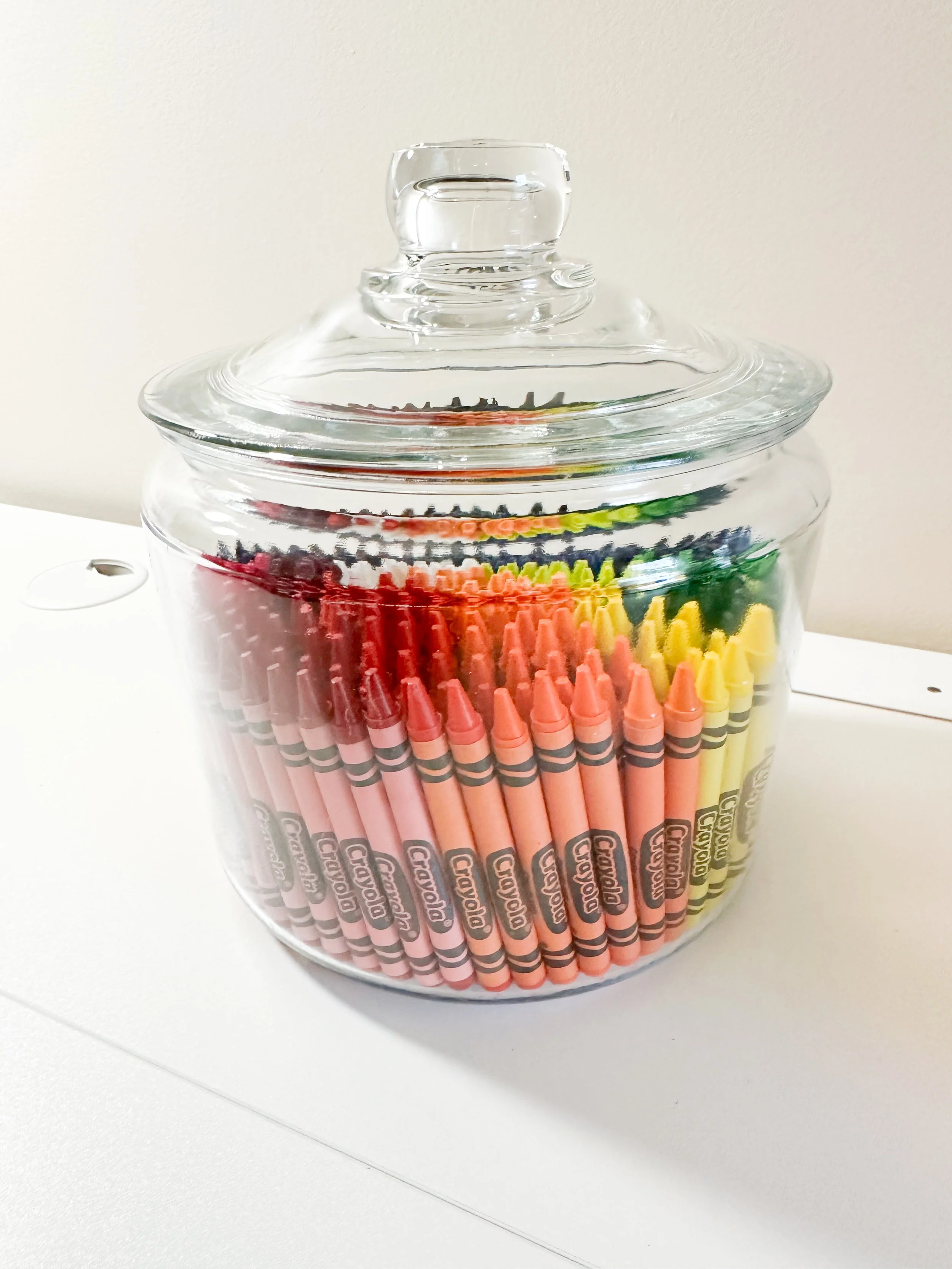 A glass jar filled with an assortment of brightly colored Crayola markers with a glass lid on top.