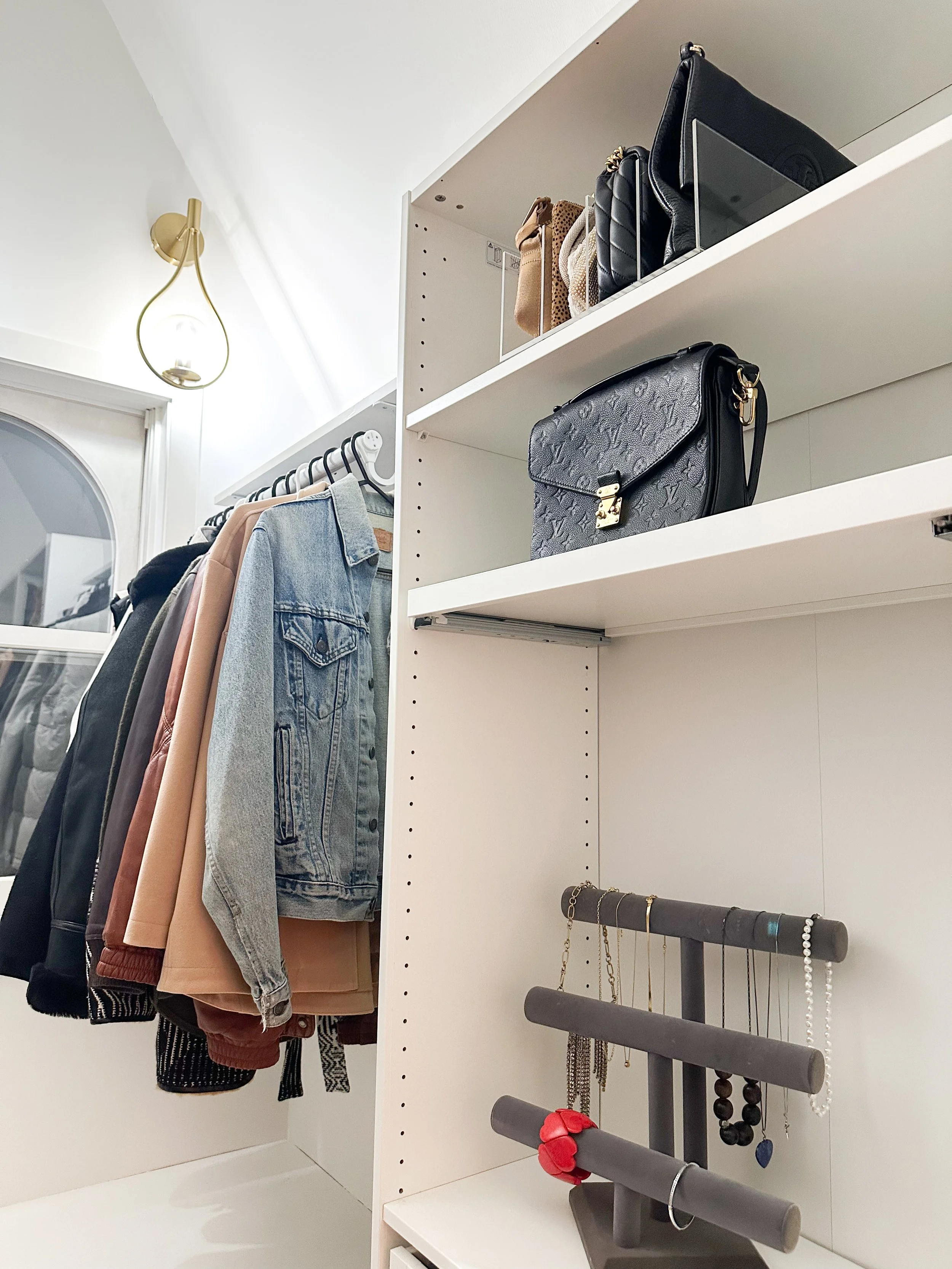Closet with hanging clothes, shelves with handbags, jewelry display, and a window.