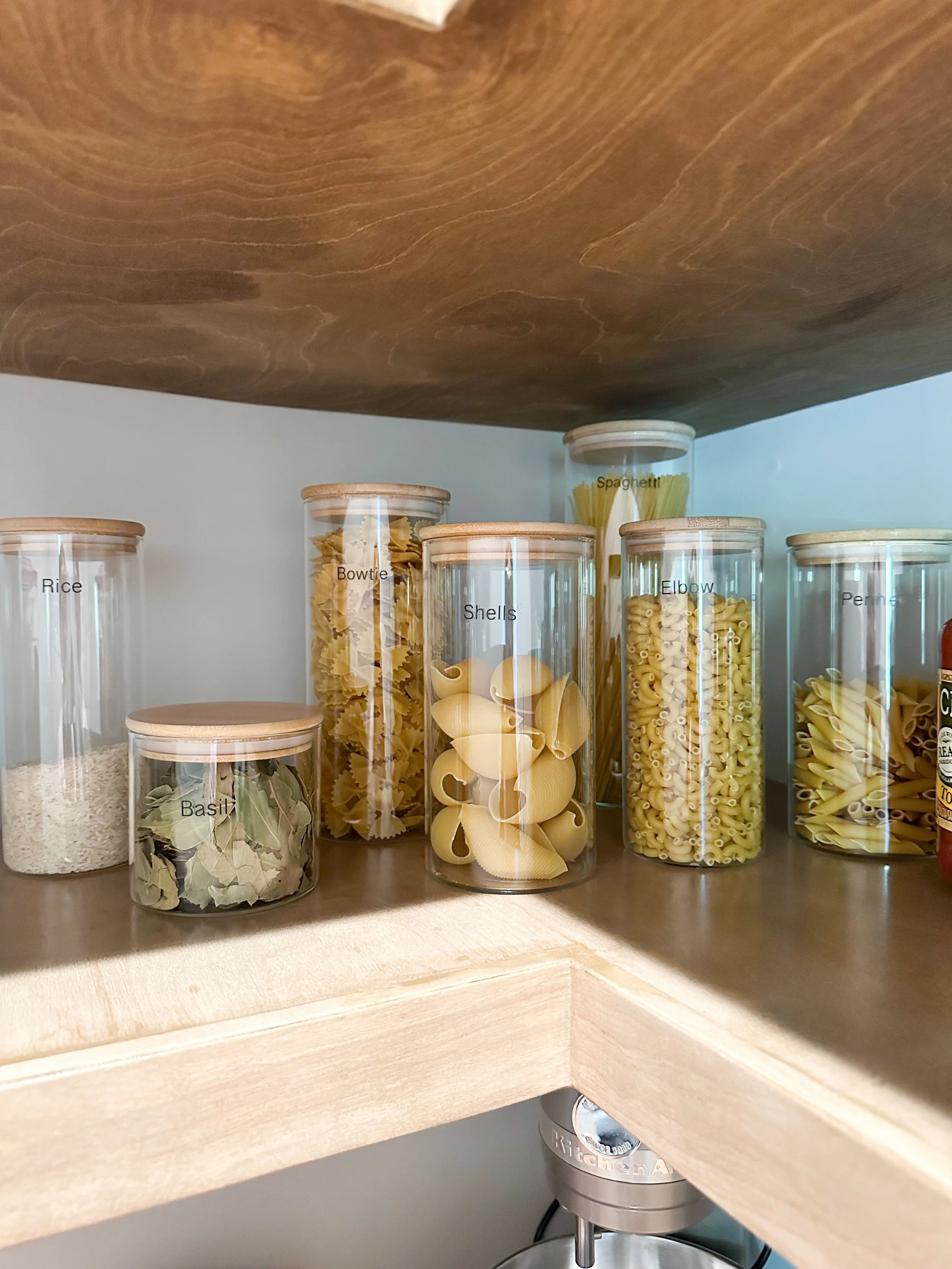 Kitchen cabinet with clear containers labeled Rice, Basil, Bowtie pasta, Shells pasta, Spaghetti, Elbow macaroni, Penne pasta. A stand mixer is visible underneath the cabinet.