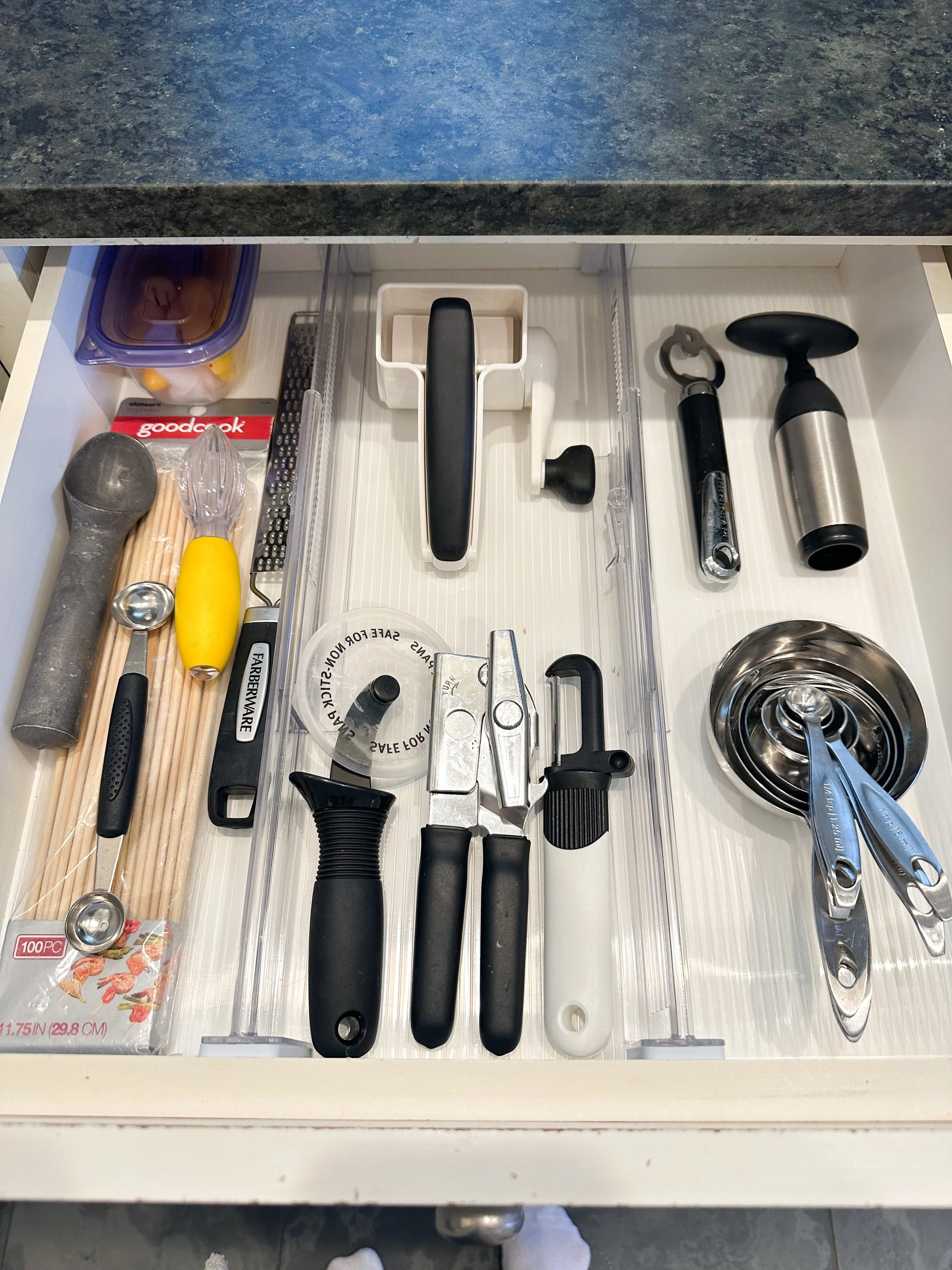 Kitchen drawer containing various utensils, including a meat mallet, dough scraper, bottle opener, measuring cups, tongs, corkscrew, and a garlic press.