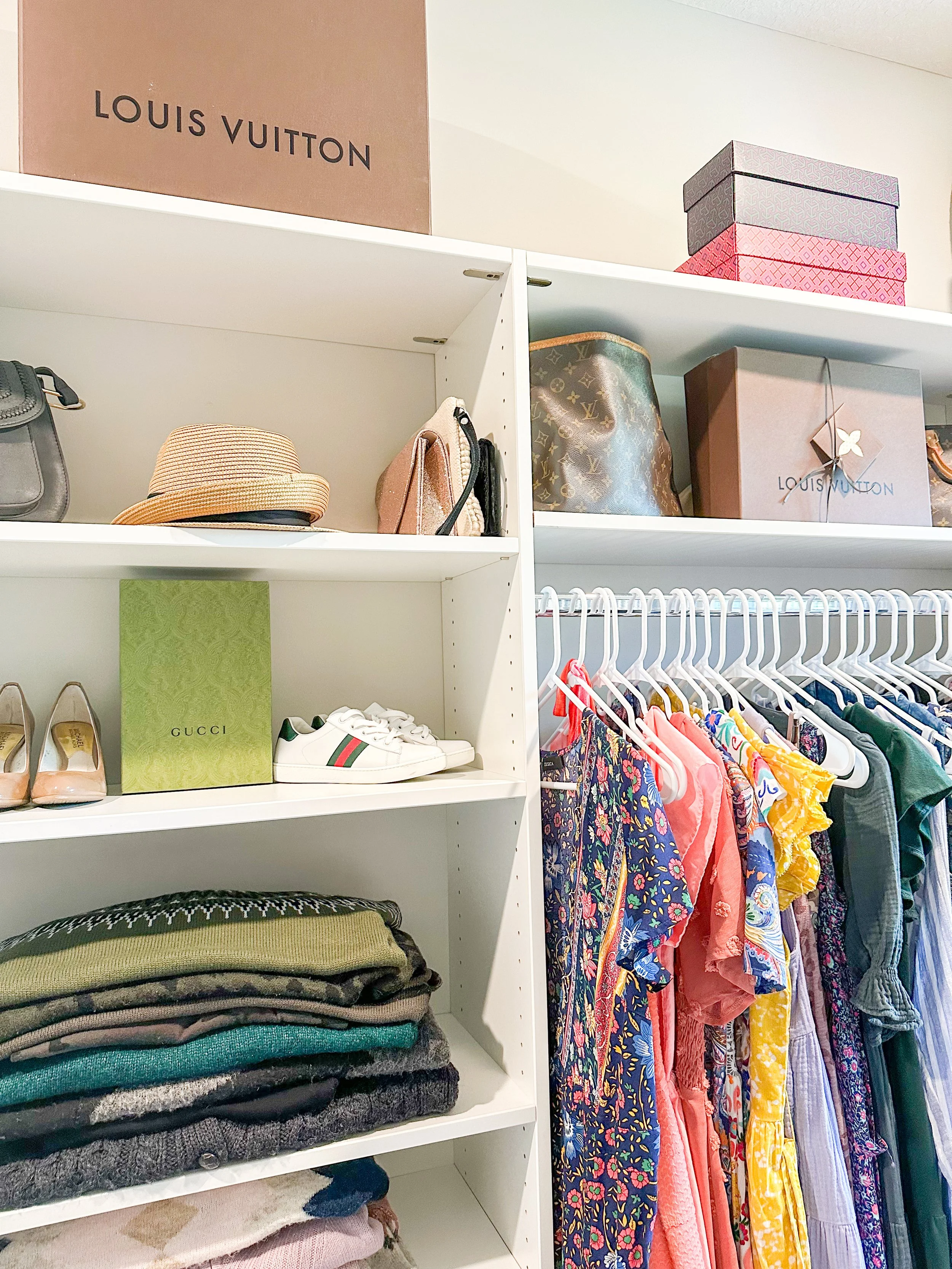 A closet shelf with designer handbags, shoeboxes, and folded clothes next to hanging colorful dresses.
