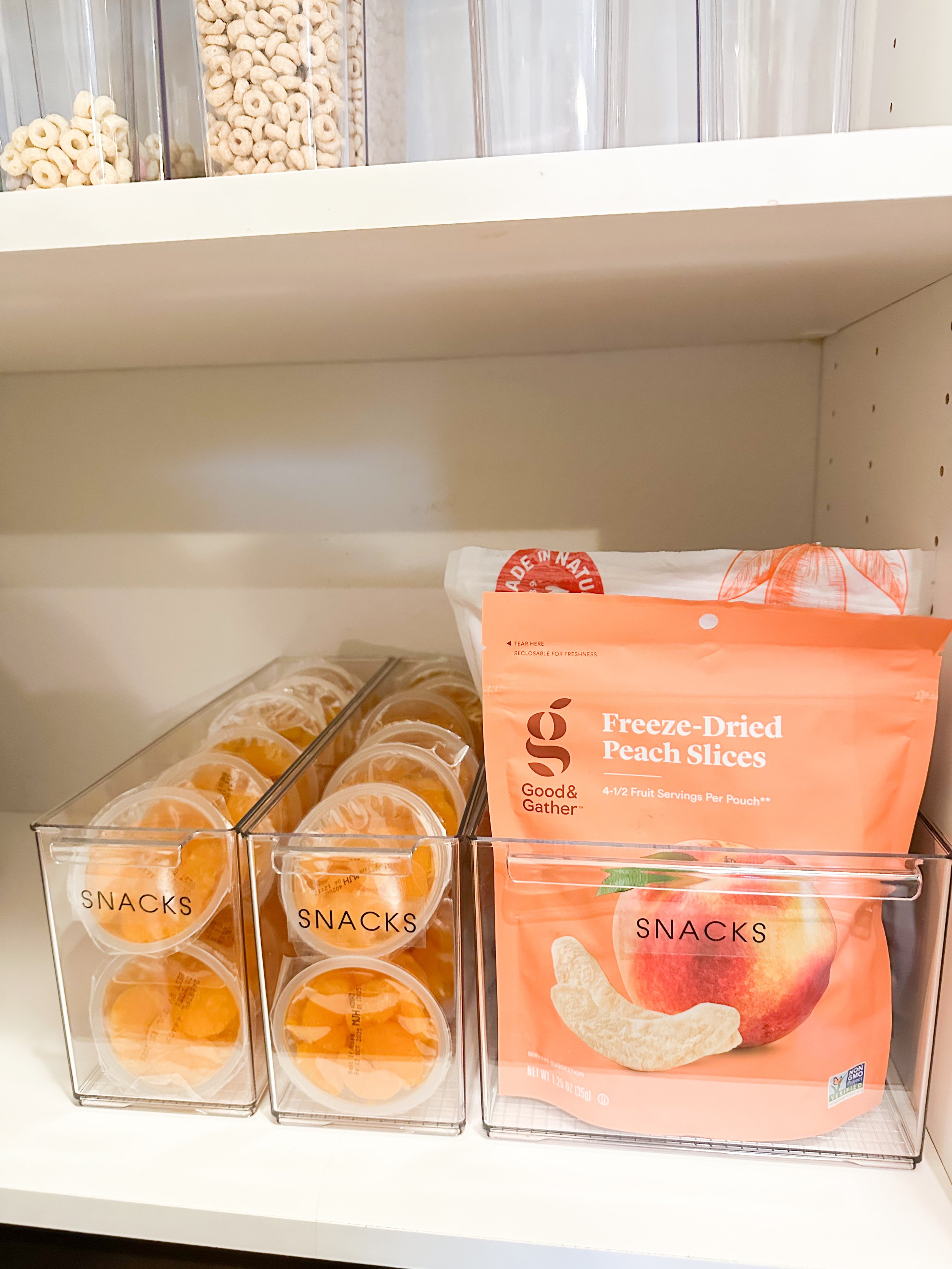 Clear plastic containers labeled 'SNACKS' filled with orange peach slices, and a bag labeled 'Freeze-Dried Peach Slices' on a white shelf.