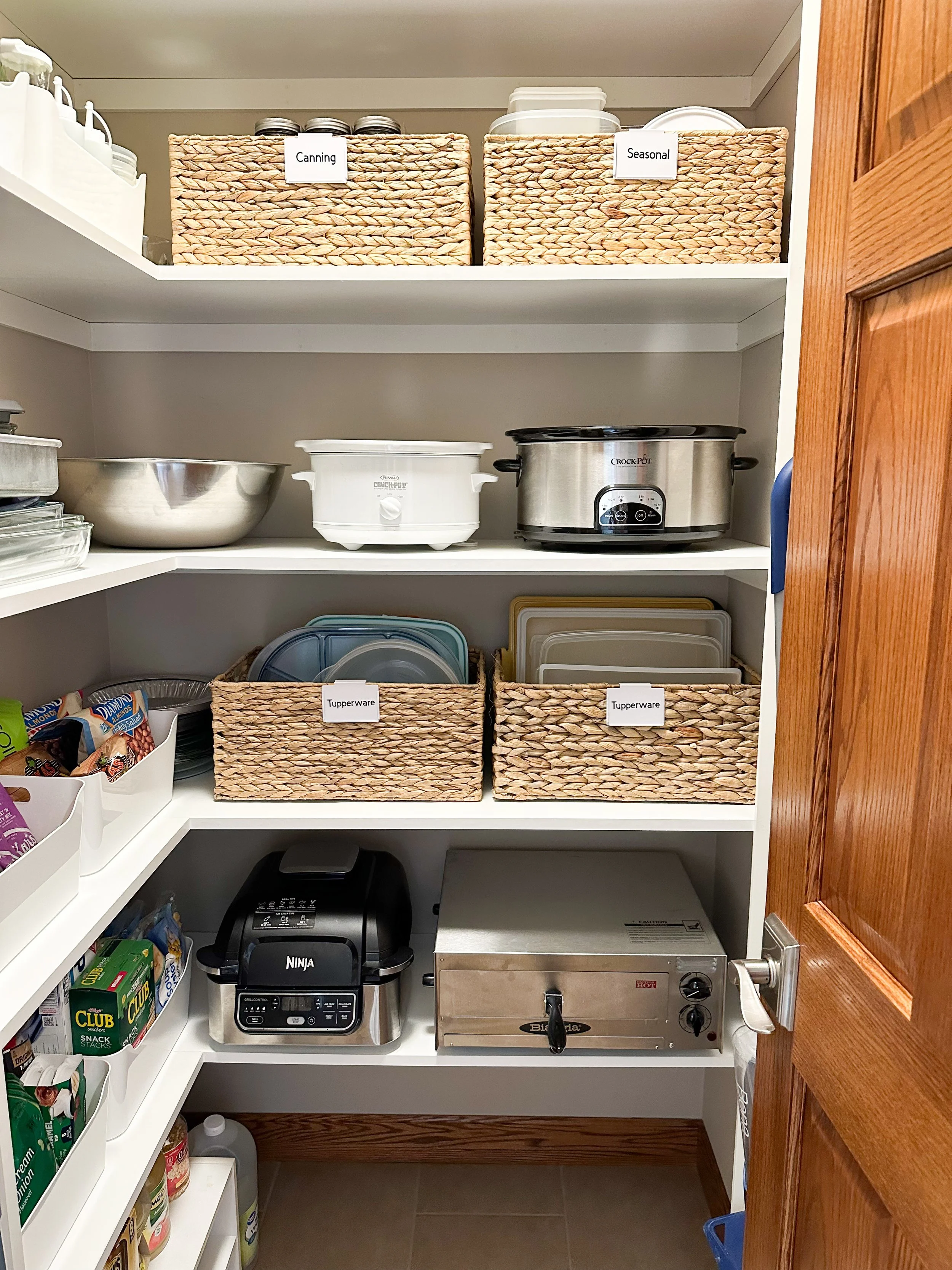 A pantry with shelves containing labeled baskets and kitchen appliances. The top shelf has wicker baskets labeled 'Canning' and 'Seasonal.' The middle shelf has a large metal mixing bowl, a white slow cooker, and a silver crockpot. The lower shelf has labeled baskets with Tupperware containers, a Ninja air fryer, and a portable oven.