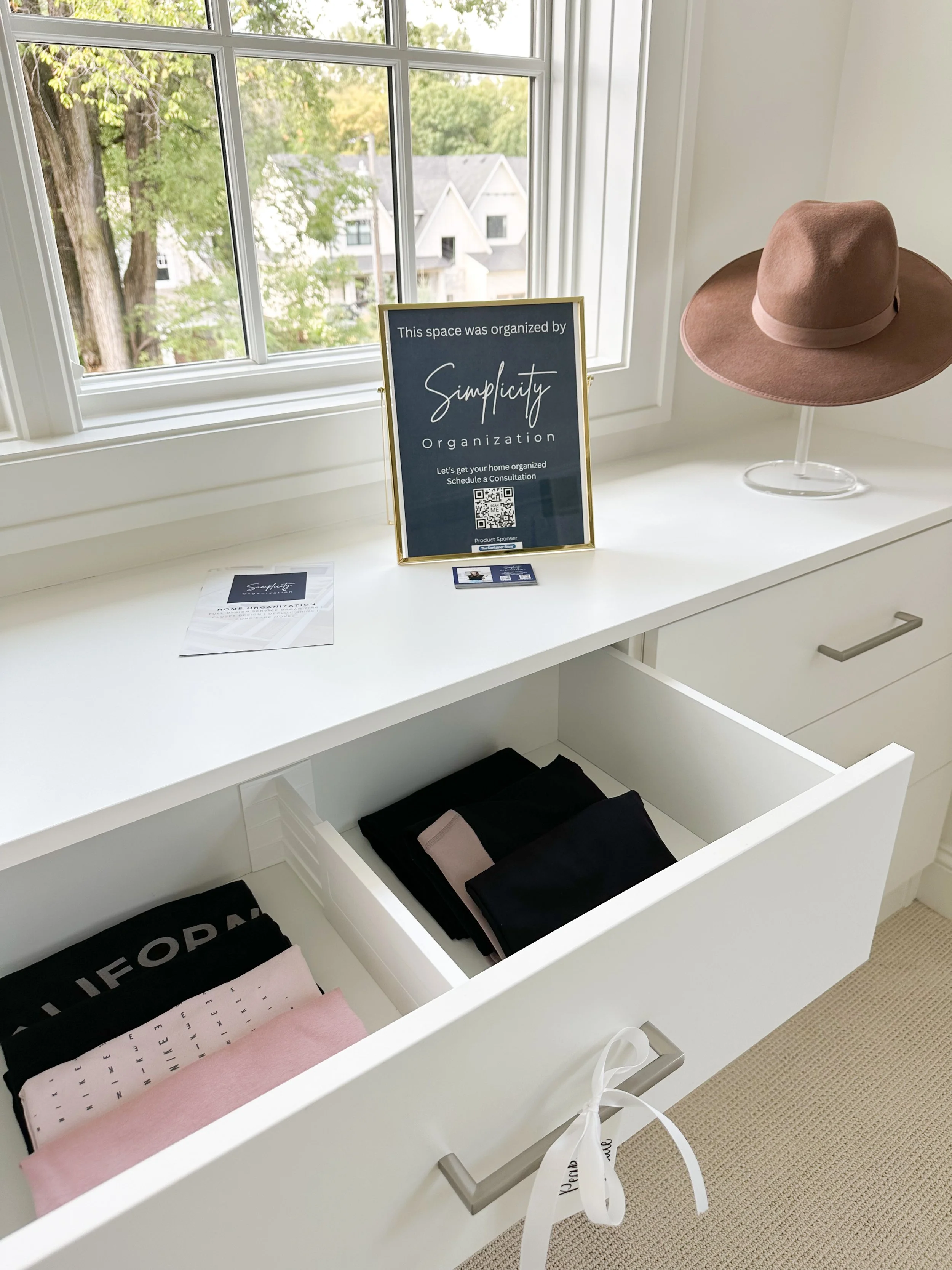 A white dresser with an open drawer containing folded clothes, a pink hat on a stand, a sign about organization, and window with trees outside.