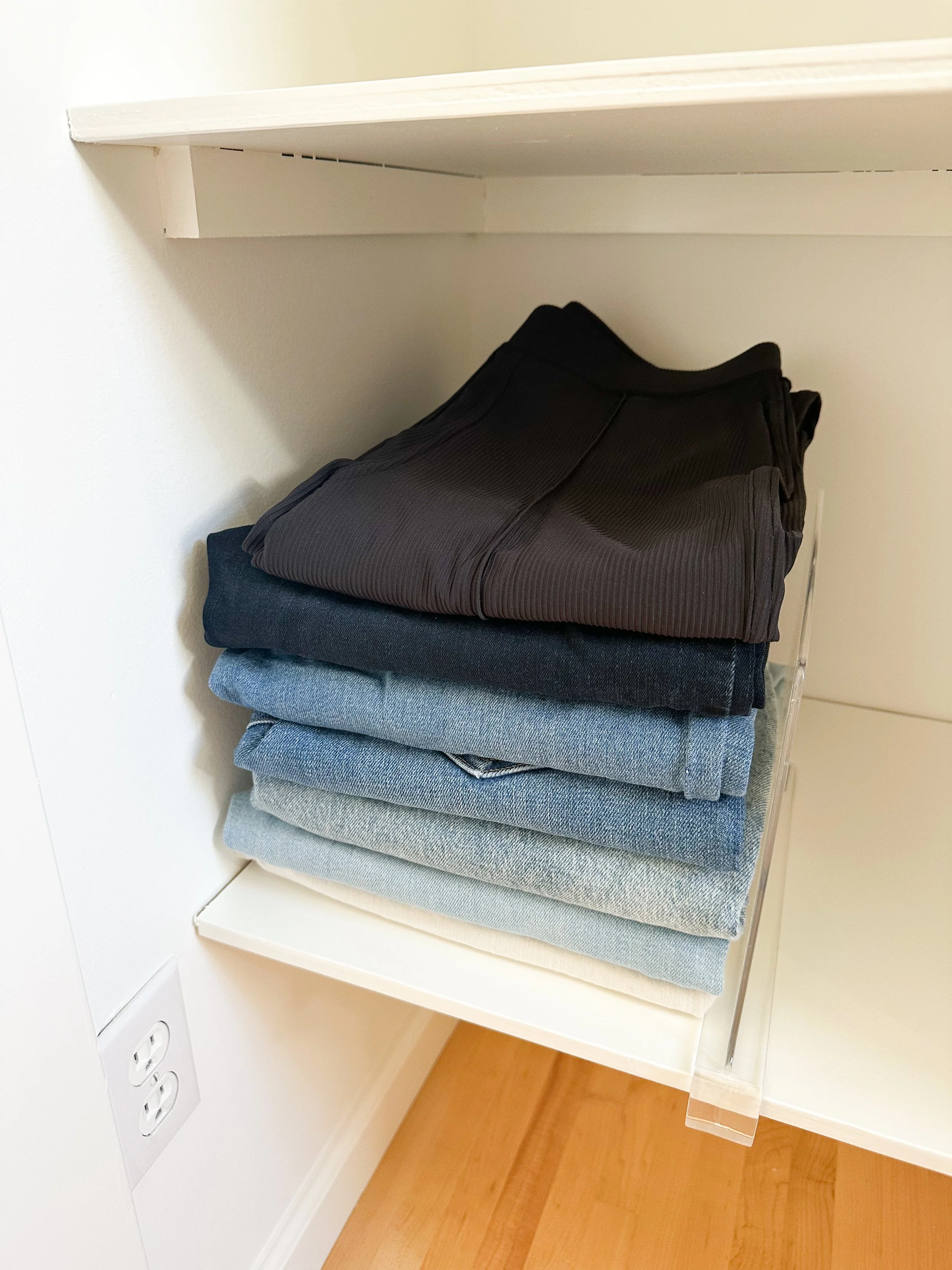 Stacked folded pants in black, dark blue, light blue, and beige on a white shelf in an indoor setting
