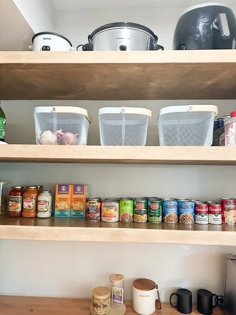 Kitchen pantry shelves with appliances on top, mesh baskets on the middle shelf containing garlic, and canned goods, jars, and coffee on the bottom shelf.