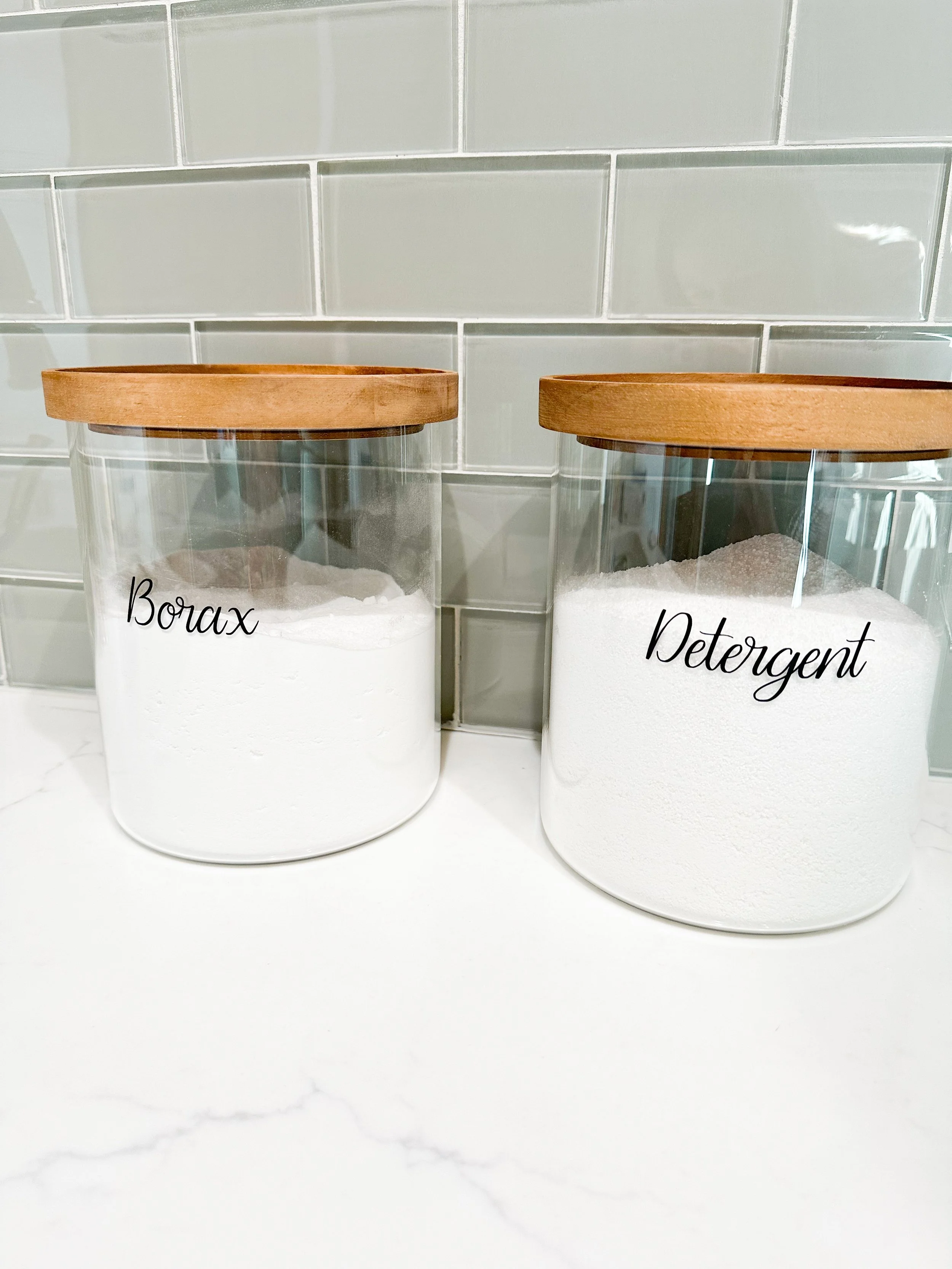 Two glass jars with wooden lids, labeled 'Borax' and 'Detergent,' containing white powders, placed on a white surface against a tiled wall.