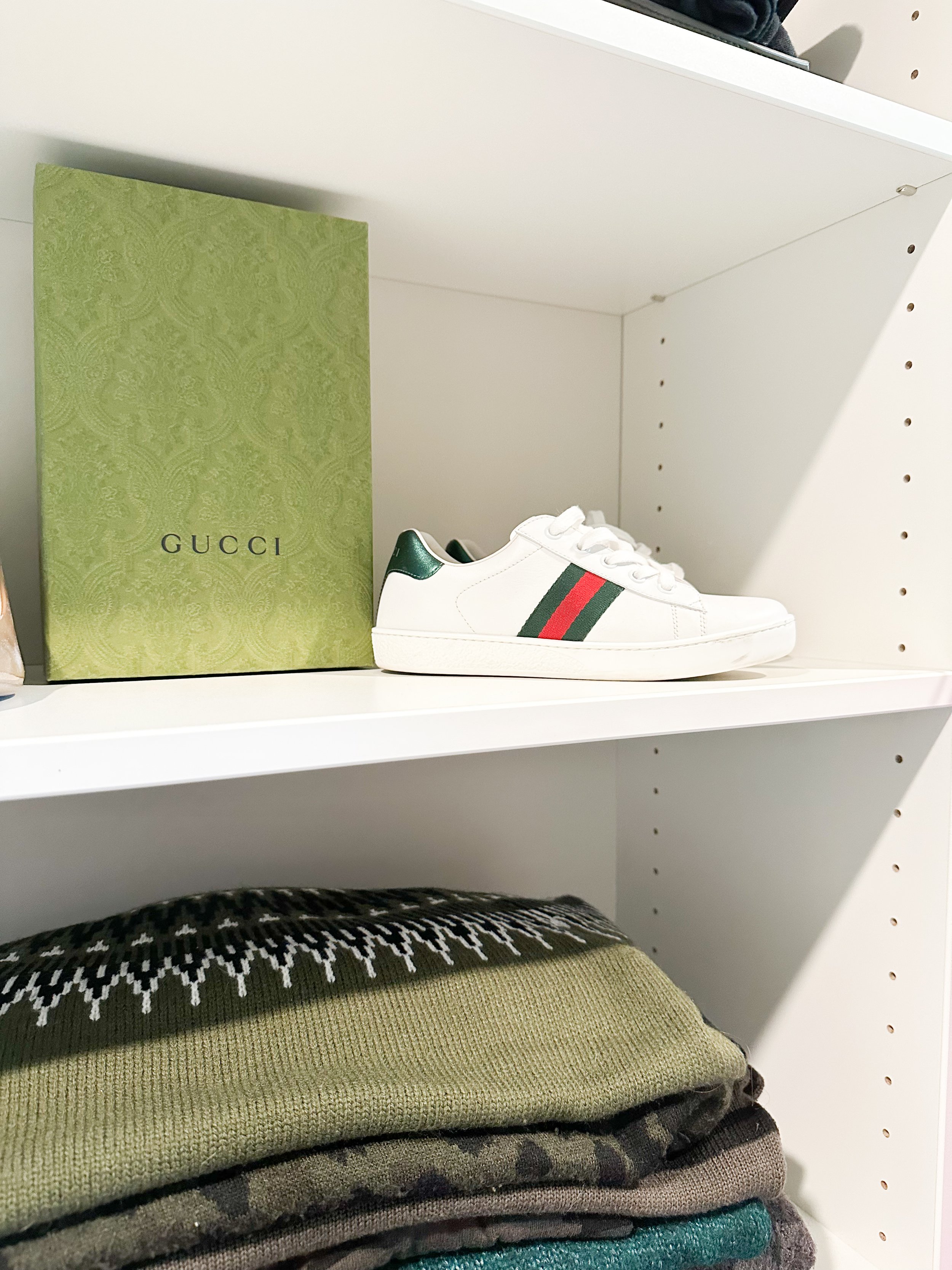 White Gucci sneaker with green and red stripes, sitting on a white shelf, with a green Gucci box next to it, and folded clothes below.