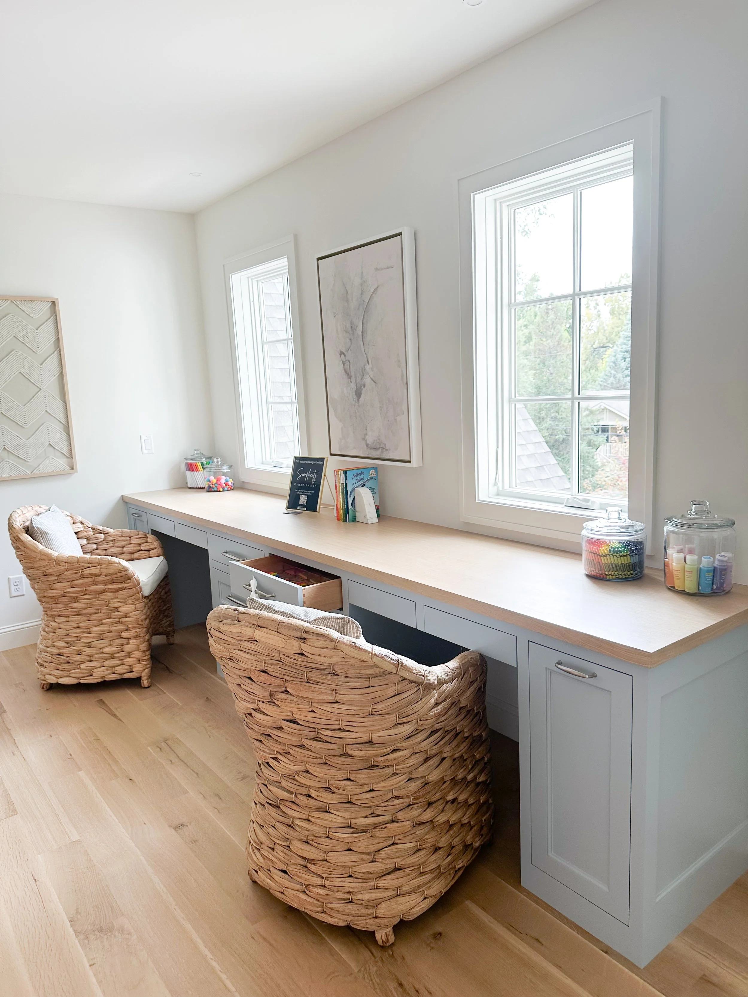 A bright room with a long wooden desk, two wicker chairs, two windows, and decorative items on the desk, including books, jars of colorful supplies, and artwork on the walls.