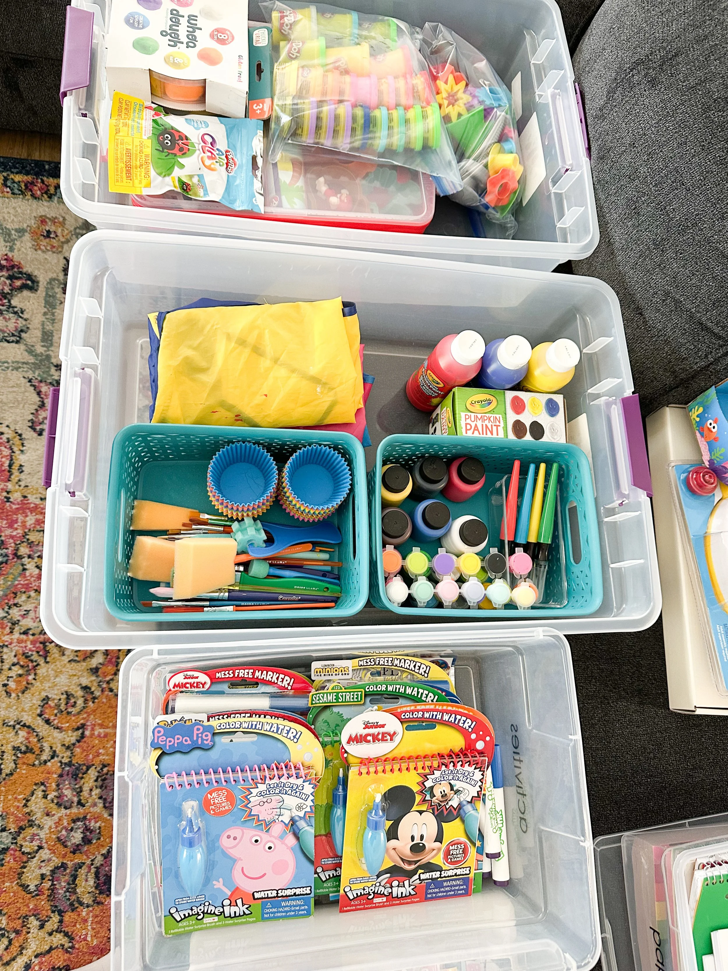 Plastic storage bins filled with children's craft supplies, including paints, markers, brushes, coloring books, and craft kits featuring Peppa Pig and Mickey Mouse.