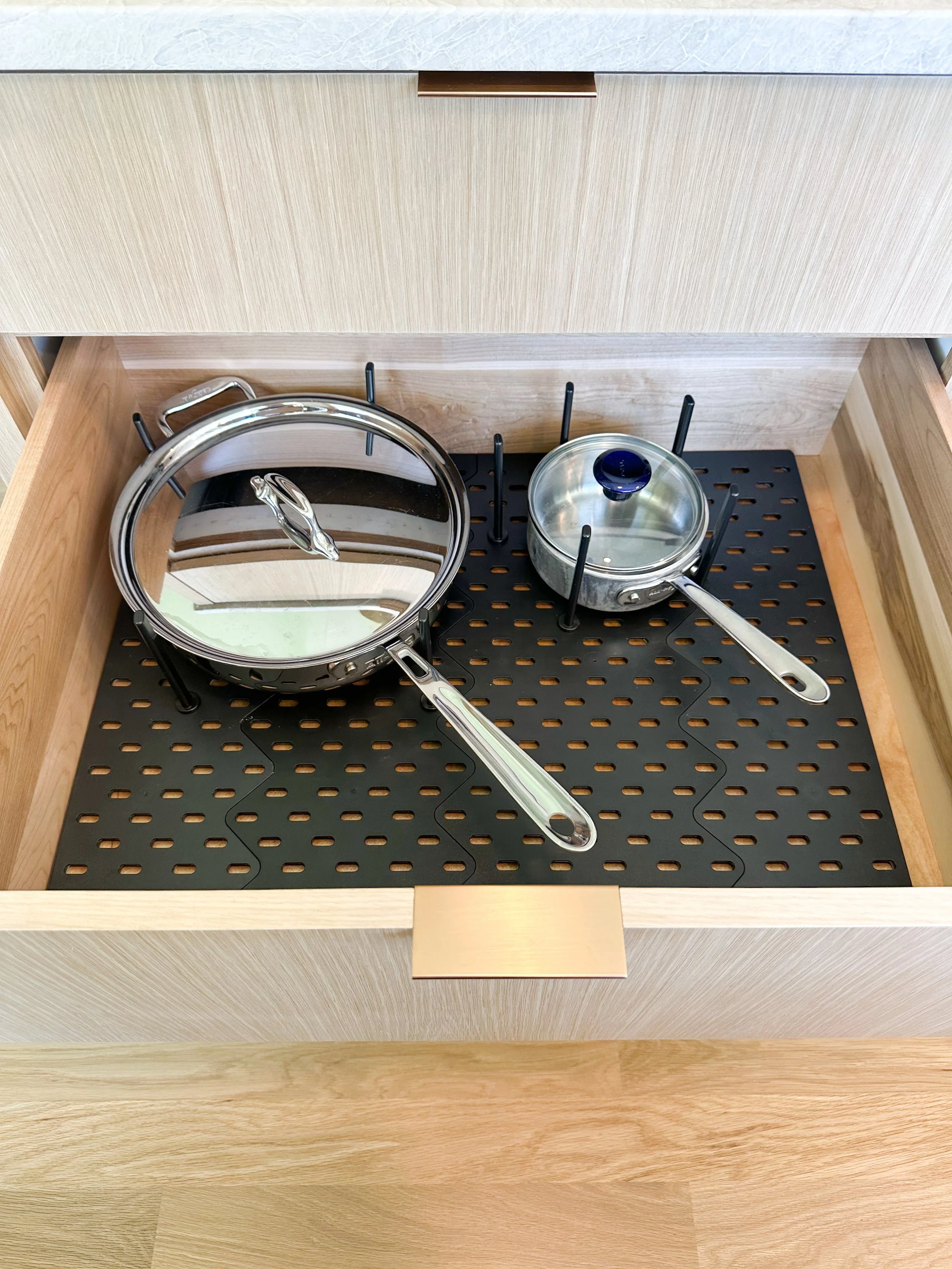Open kitchen drawer with two stainless steel pots with glass lids, one large and one small, inside a kitchen drawer with a black mat at the bottom.