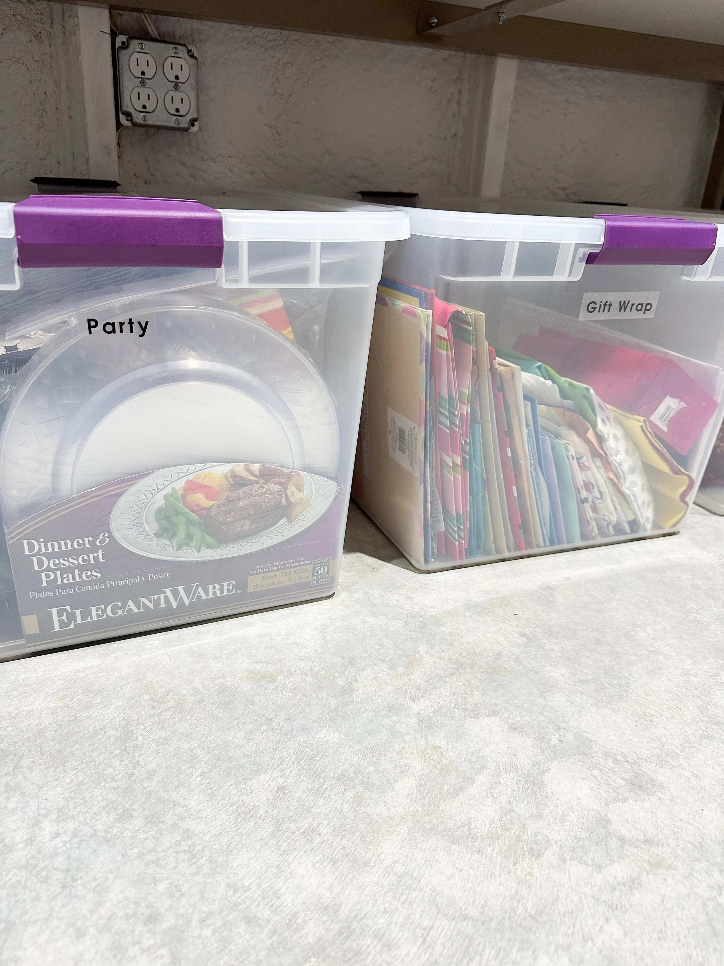 Two clear plastic storage containers with purple lids containing disposable dinnerware and party supplies, labeled 'Party' and 'Gift Wrap' on a light-colored surface against a wall with an electrical outlet.