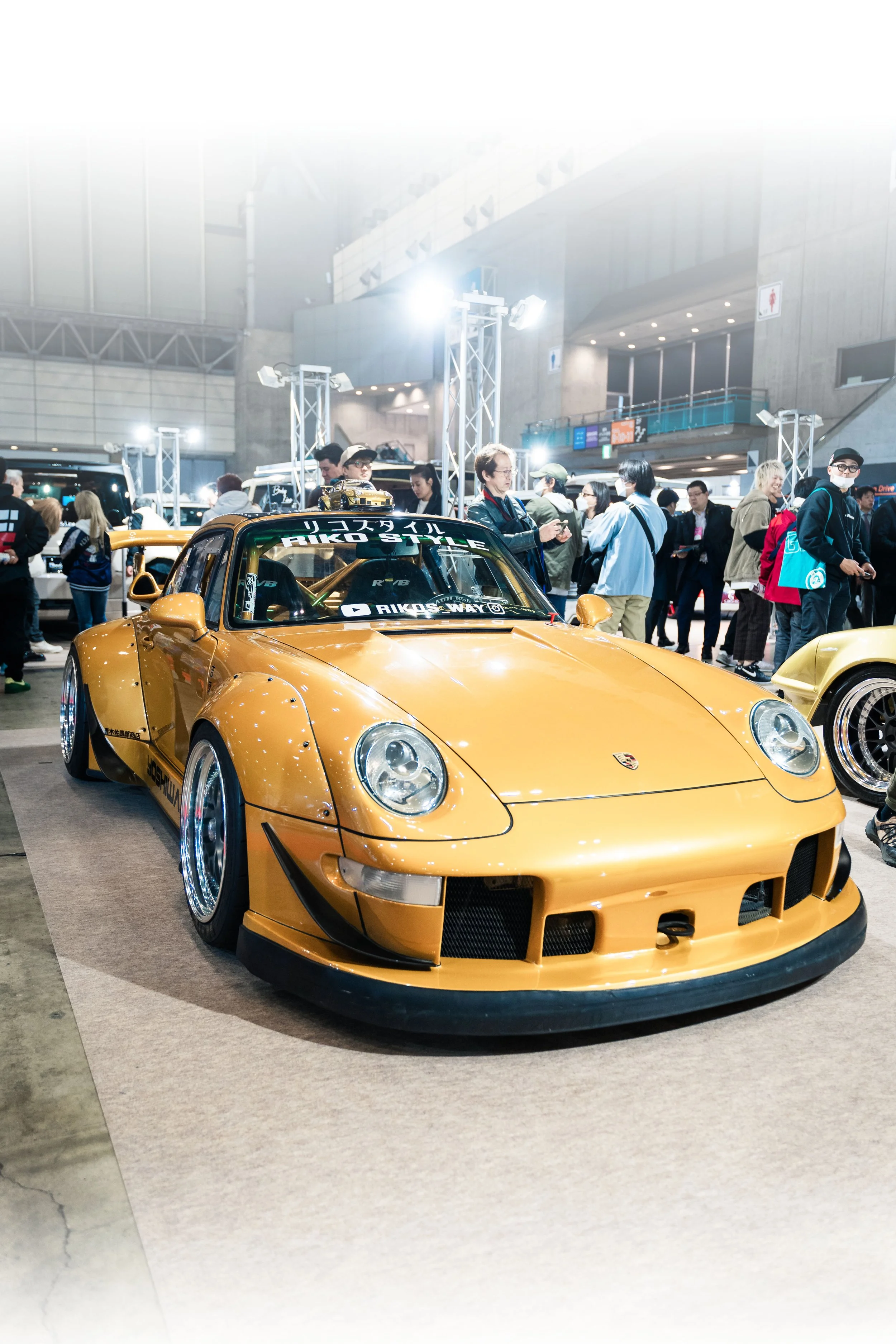 Gold Porsche sports car on display at an indoor auto show with people in the background.