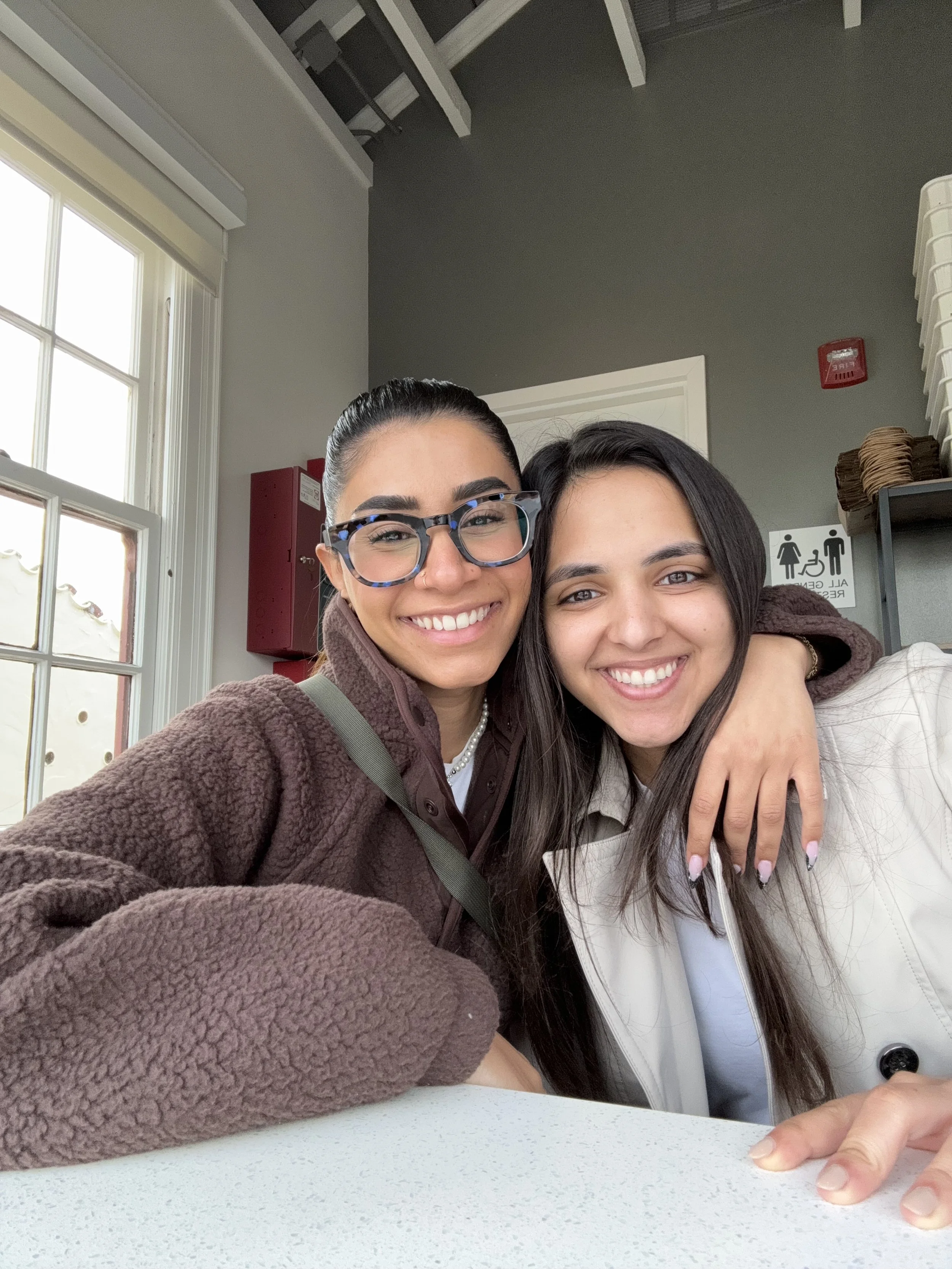 Two women smiling and hugging at a table inside a building with gray walls and a window in the background.