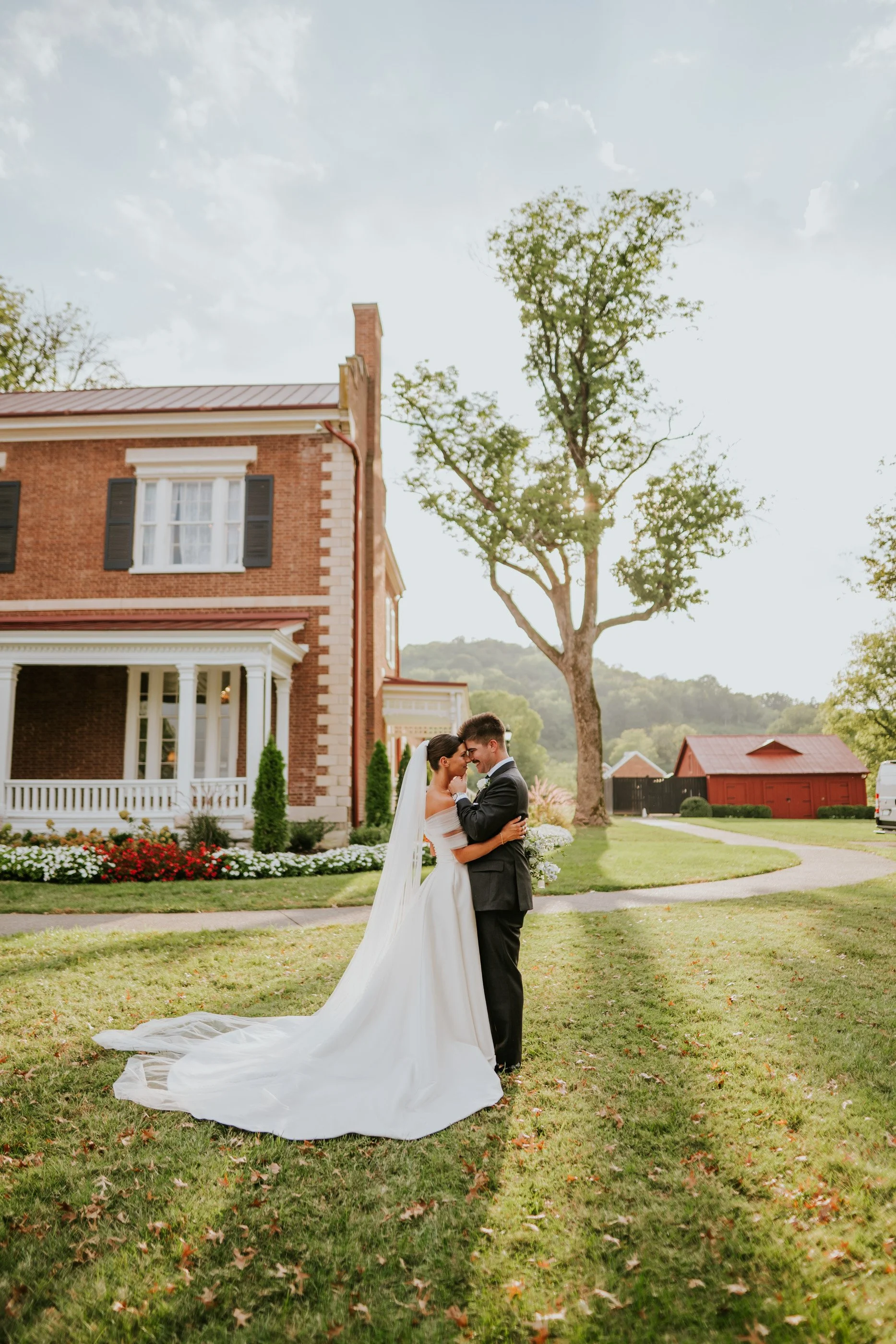 A bride in a white wedding dress and a groom in a black tuxedo stand close together on a lawn, sharing a romantic moment during sunset, with a multi-story brick house and trees in the background.