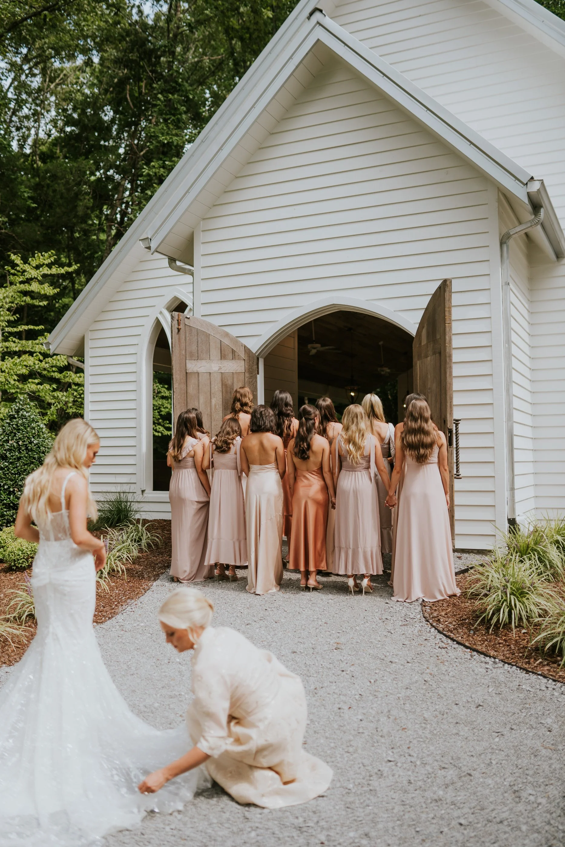 A group of women in bridesmaid dresses are entering a white chapel with wooden doors, with two women in wedding dresses in the foreground.