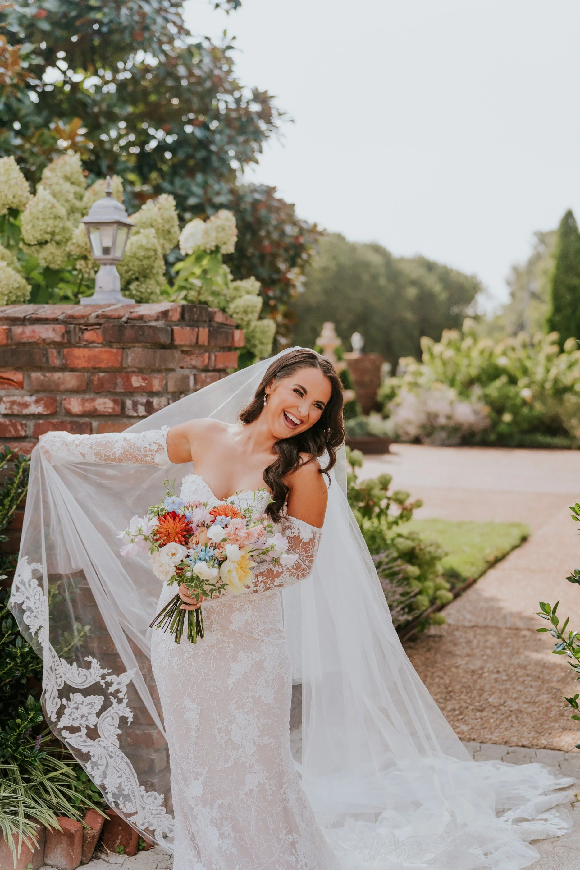 Bride in a lace wedding dress and veil holding a colorful bouquet, smiling outdoors in a garden setting with greenery and brick accents.