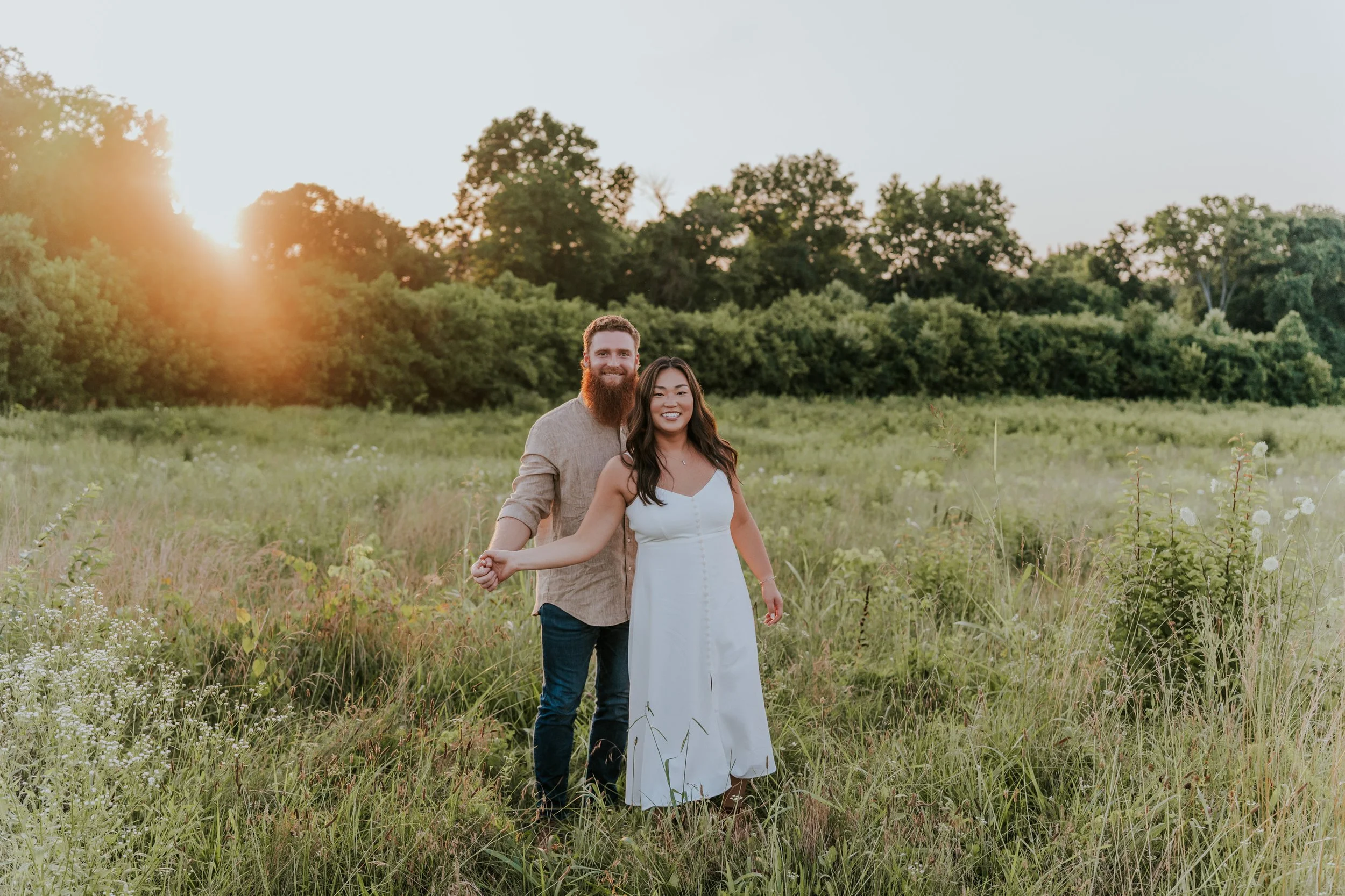 A happy couple standing hand in hand in a grassy field at sunset, smiling at the camera.
