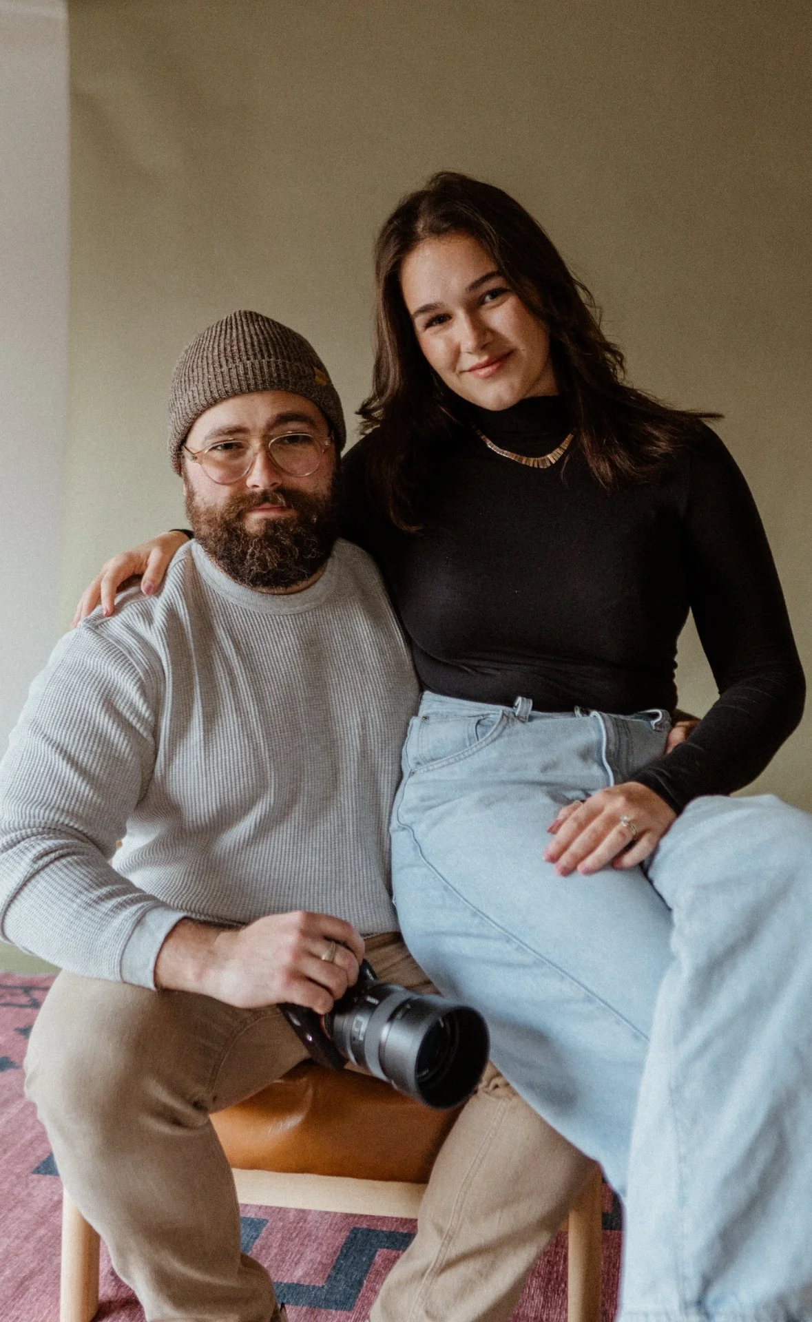 A man with glasses, a beanie, and a beard sitting on a chair, holding a camera, with a woman sitting on his lap, both smiling at the camera.