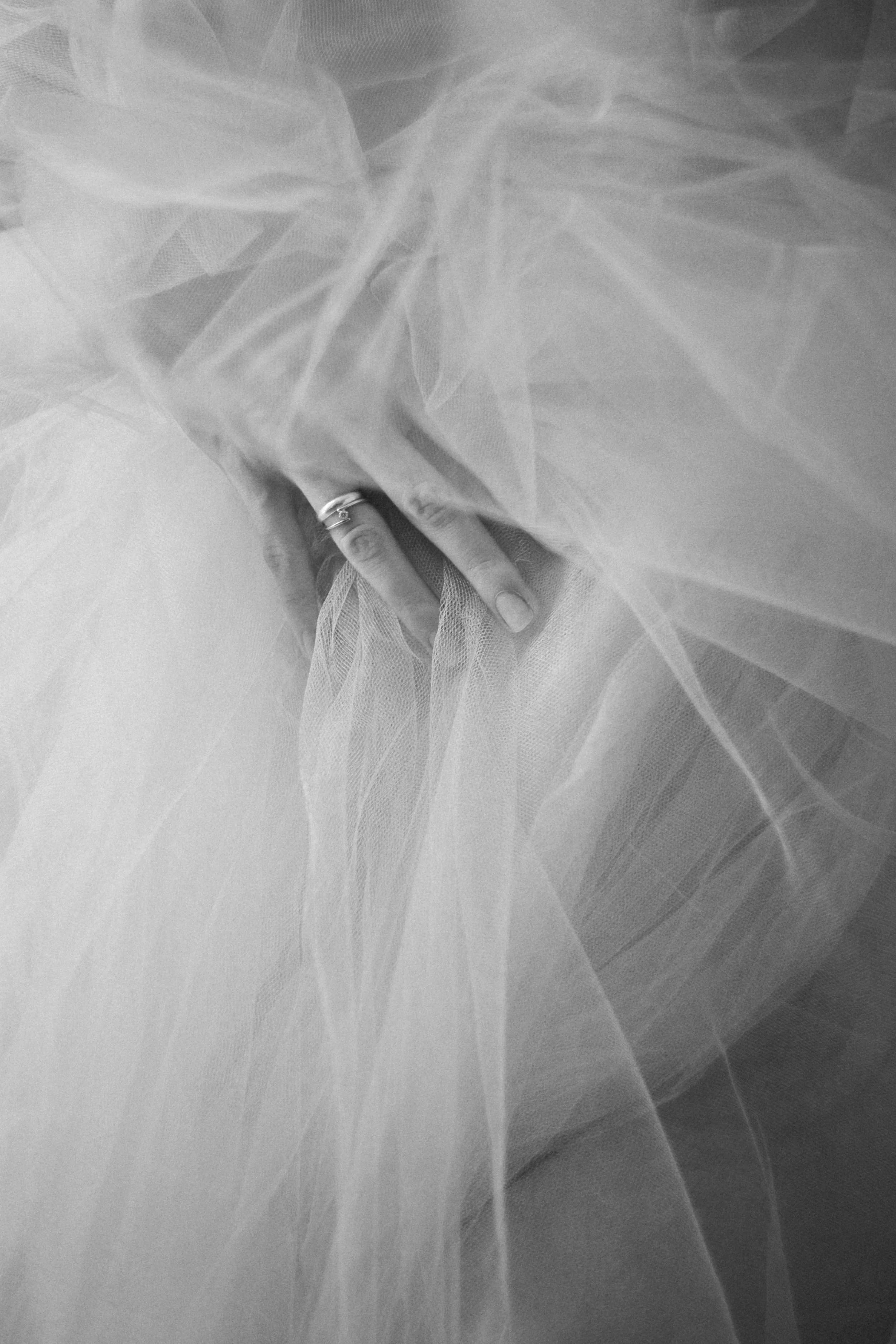 A hand with a ring on the finger is partially covered by soft, flowing, sheer fabric that creates an ethereal effect in a black-and-white photograph.