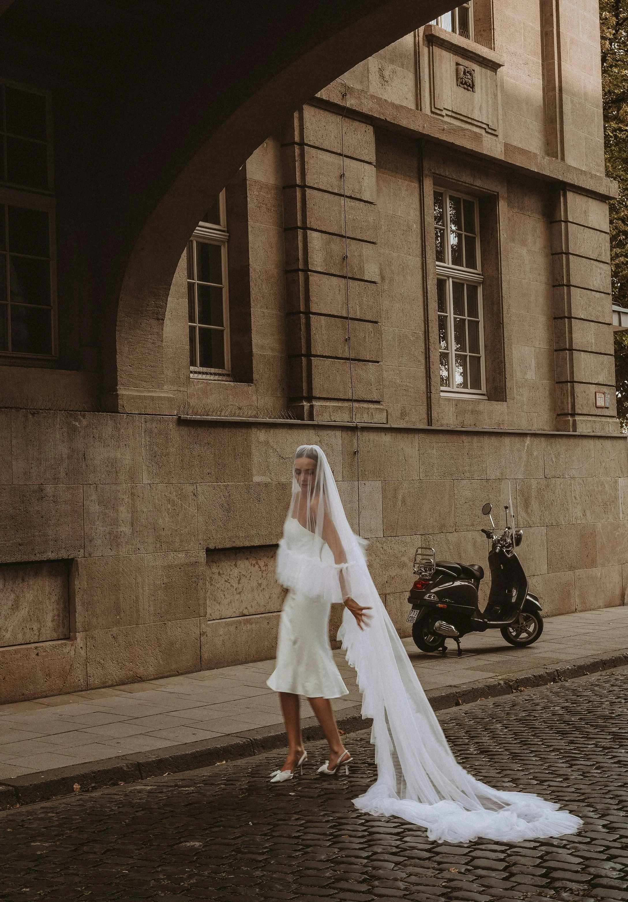A woman in a white dress and veil standing on a cobblestone street next to a scooter, in front of a historic stone building with large windows.