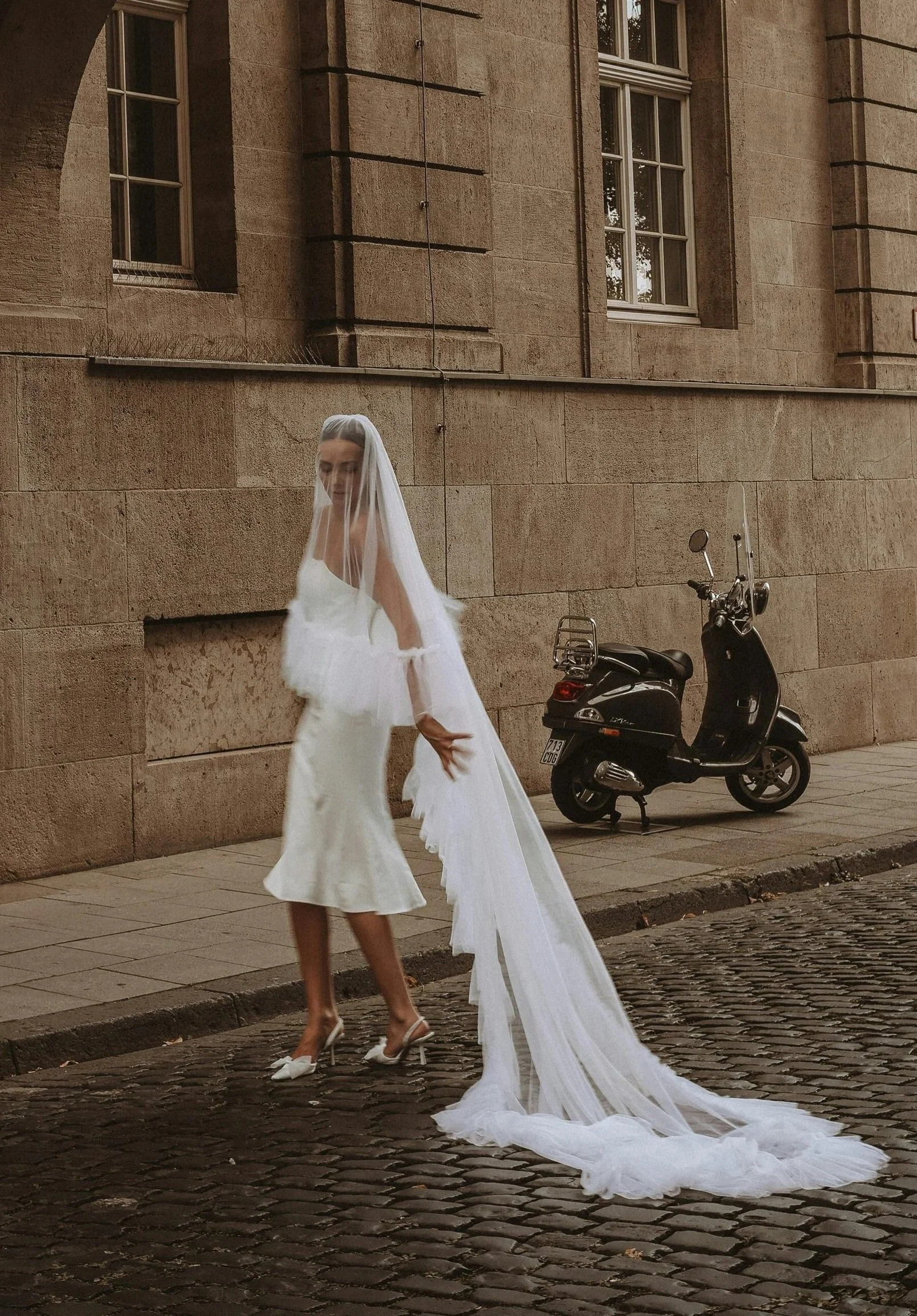 A woman in a white wedding dress and veil standing on a cobblestone street, with a black scooter parked against a stone building in the background.