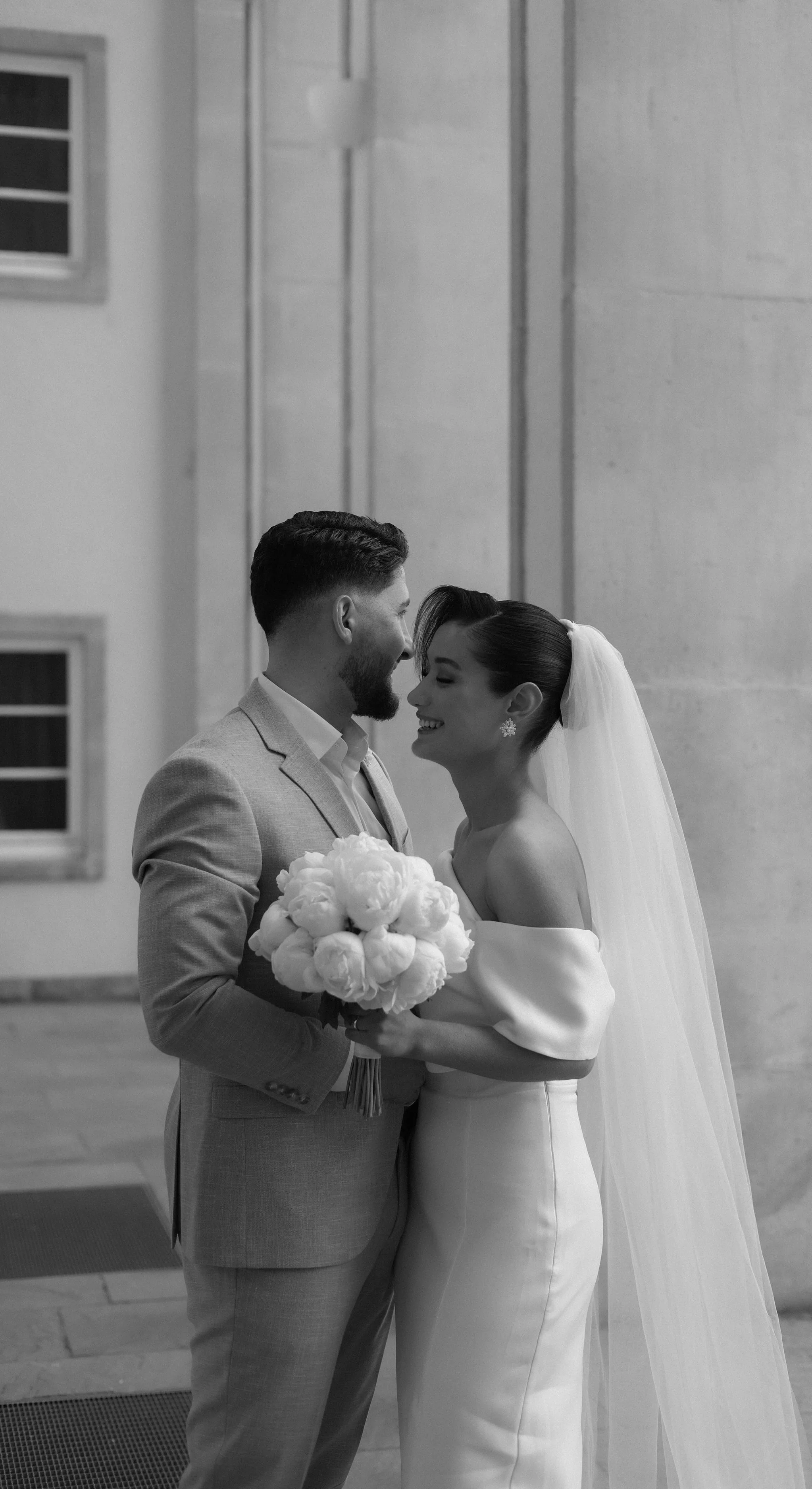 A black and white photo of a bride and groom standing close together, smiling, with the bride holding a bouquet of flowers and wearing a veil, indoors.