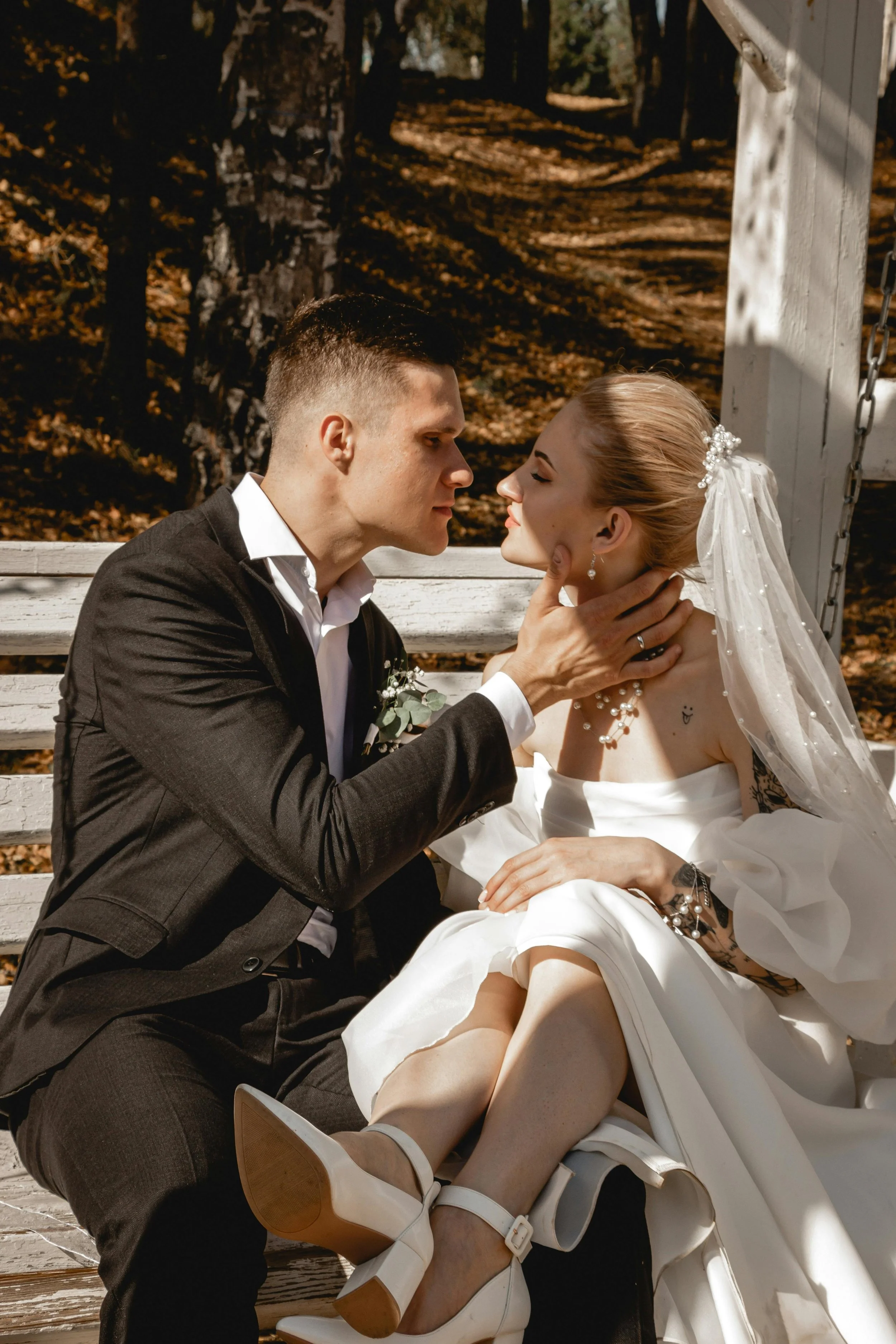 A bride and groom sitting on a park bench during their wedding, with the groom gently holding the bride's chin as they look into each other's eyes, surrounded by autumn trees.