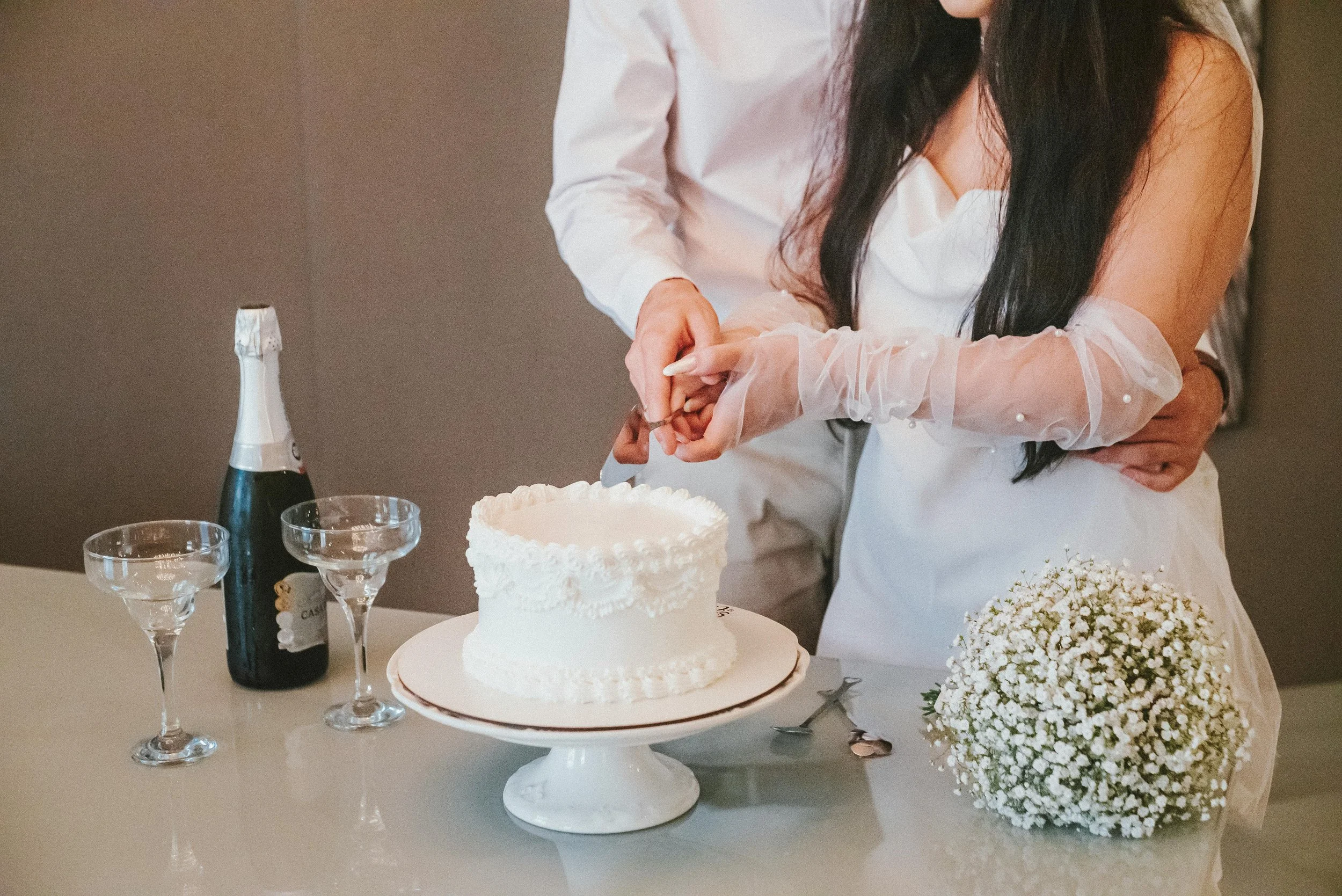 Couple cutting wedding cake with champagne, glasses, flowers, and celebration items on table.