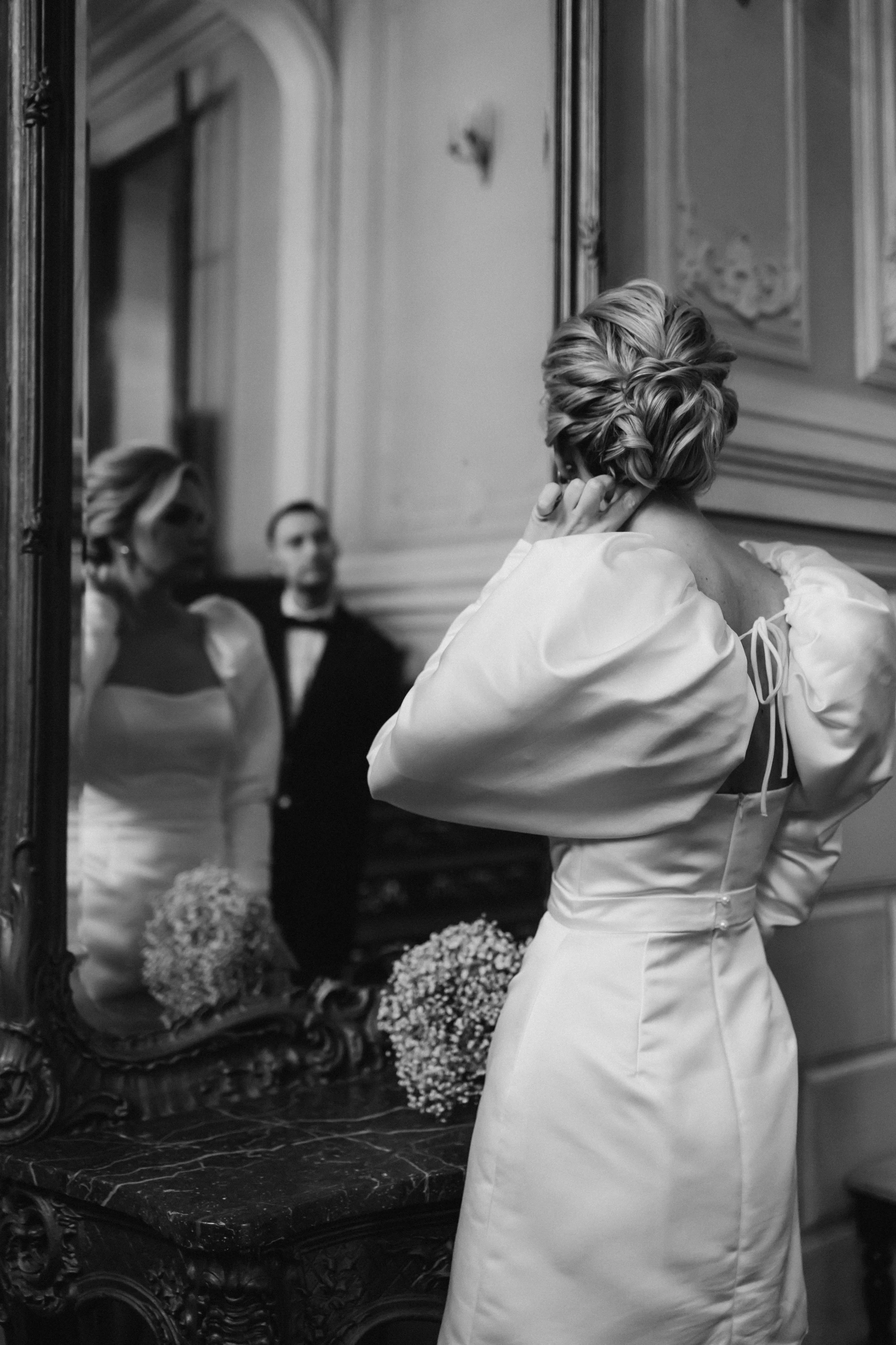 A woman in an elegant dress looks at her reflection in a mirror, adjusting her hair before a wedding or formal event, with another woman and a man in the background.