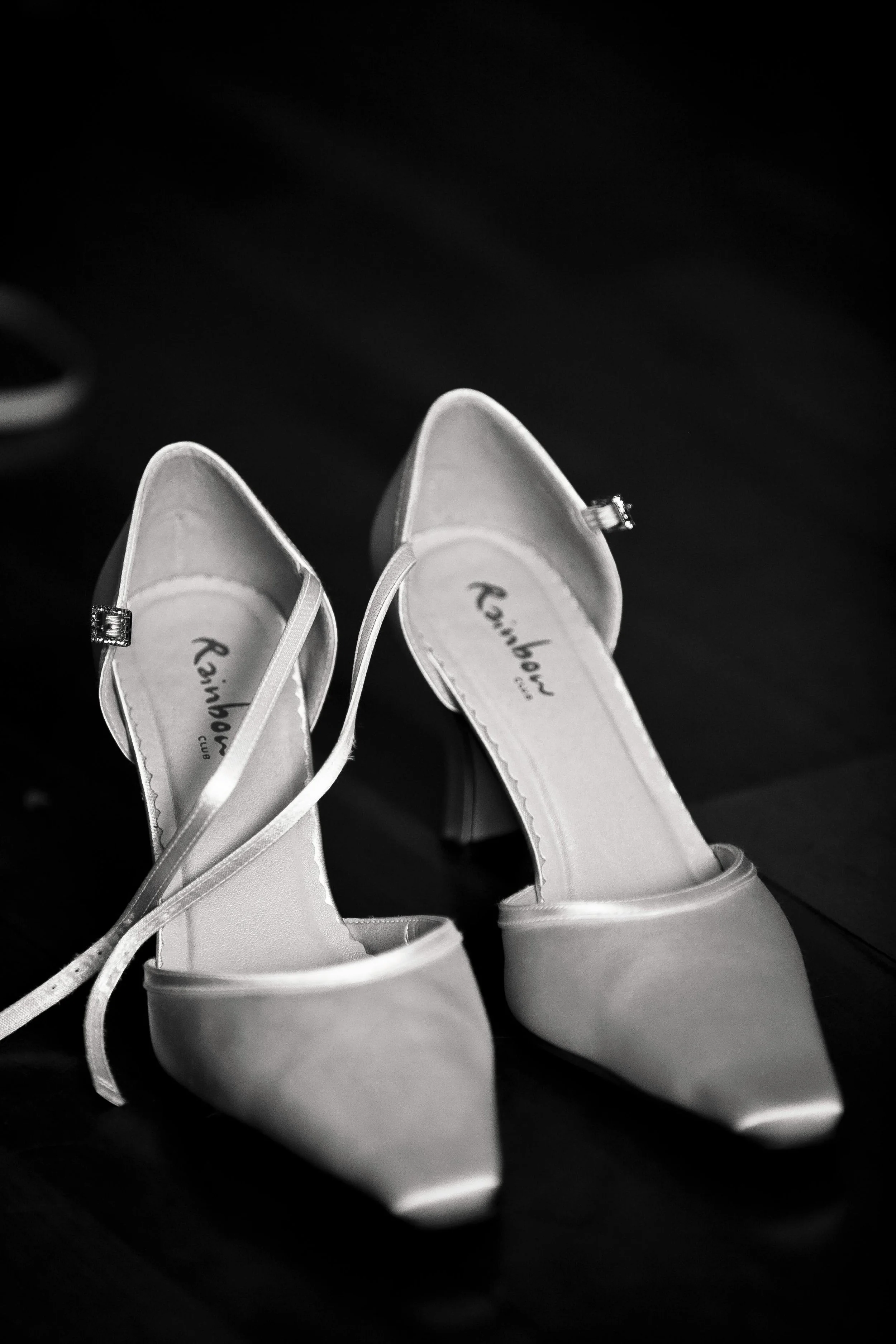 A pair of satin wedding heels with pointed toes and ankle straps, placed on a dark surface.