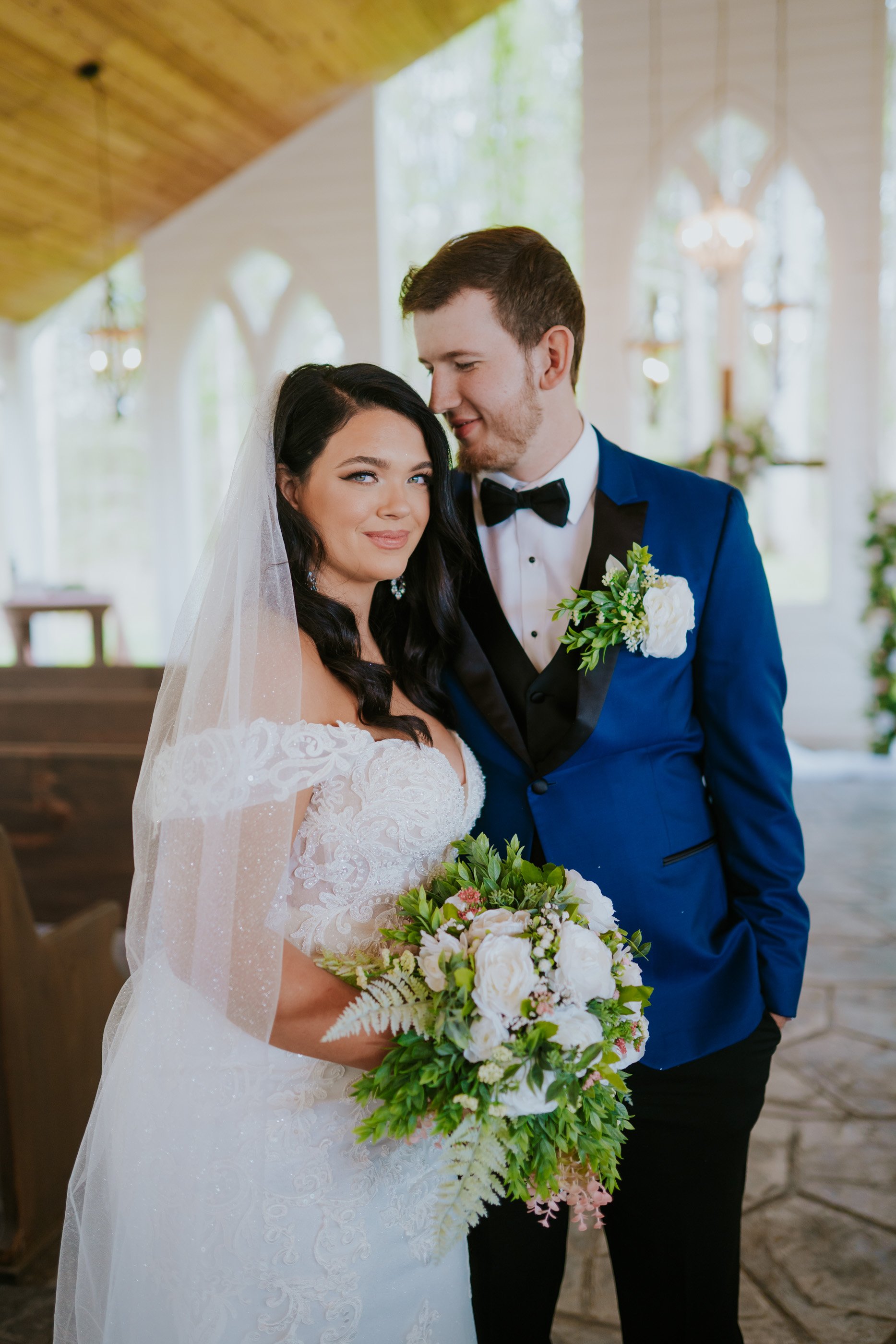 Bride and groom standing inside a church with floral arrangements, dressed in wedding attire, with the groom in a blue tuxedo and the bride holding a bouquet of white and green flowers.