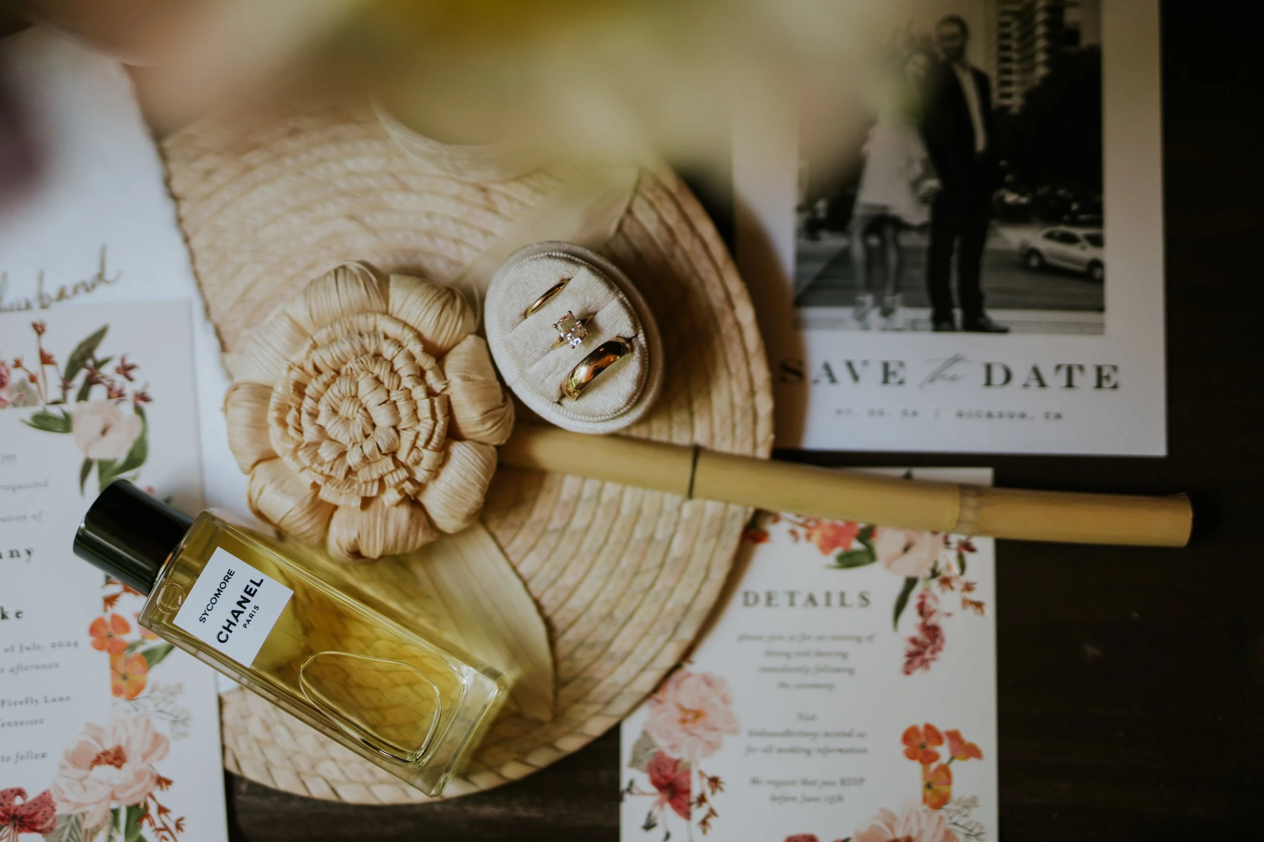 Wedding rings in a ring box, Chanel perfume bottle, a silk flower, a floral wedding invitation, a save the date card, and a photo of a couple on a wedding day.