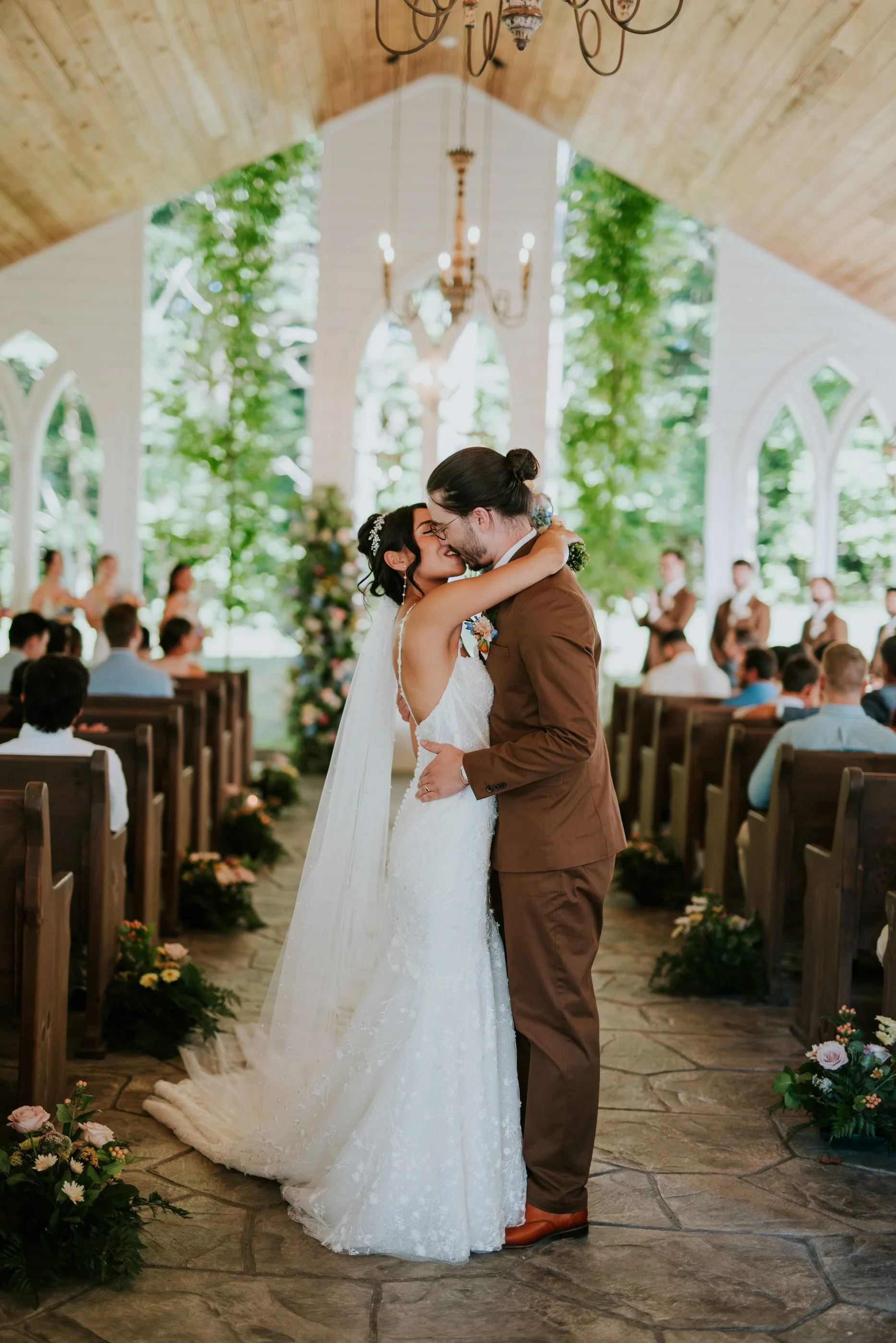 A bride and groom sharing a kiss during their wedding ceremony inside a chapel decorated with flowers and greenery, with guests seated on wooden pews in the background.