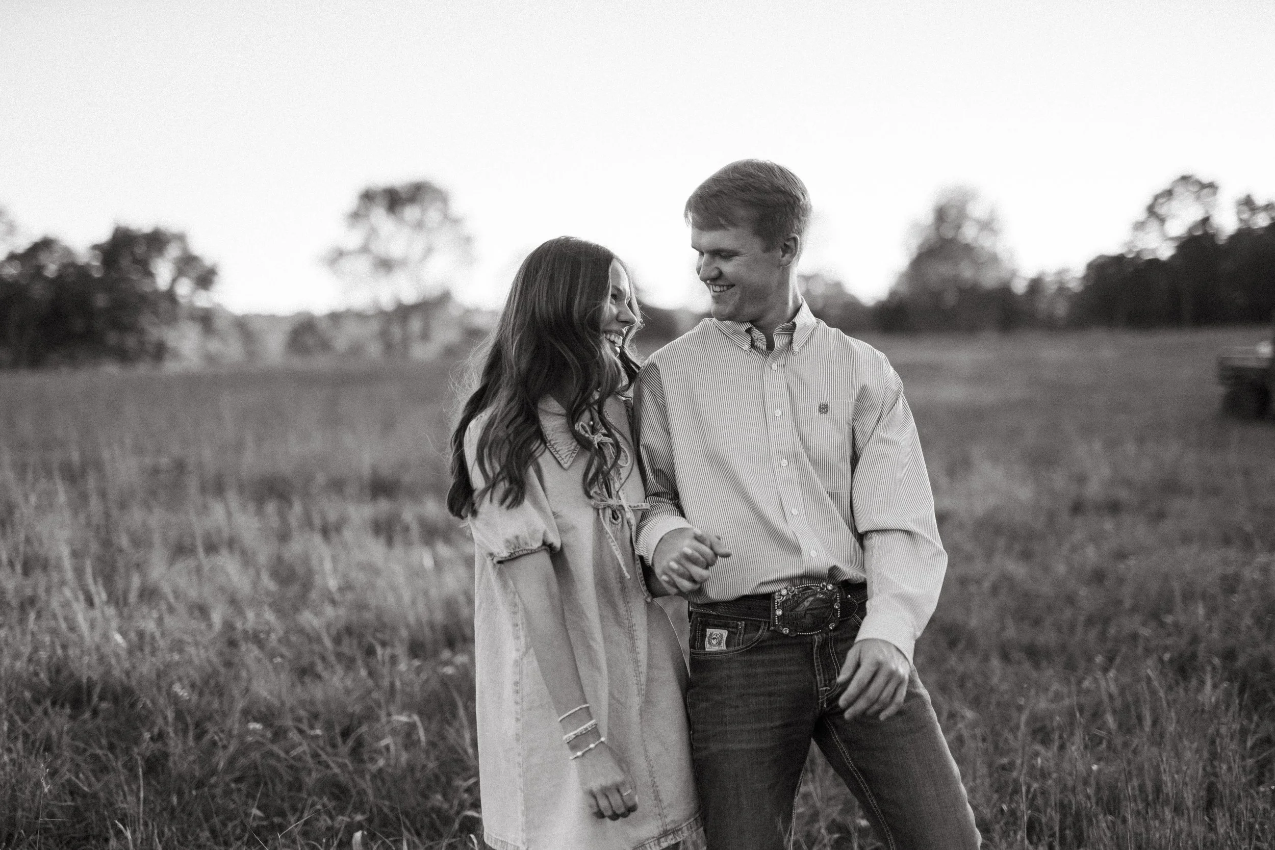 A young couple, smiling and holding hands, walking in a field with trees in the background.