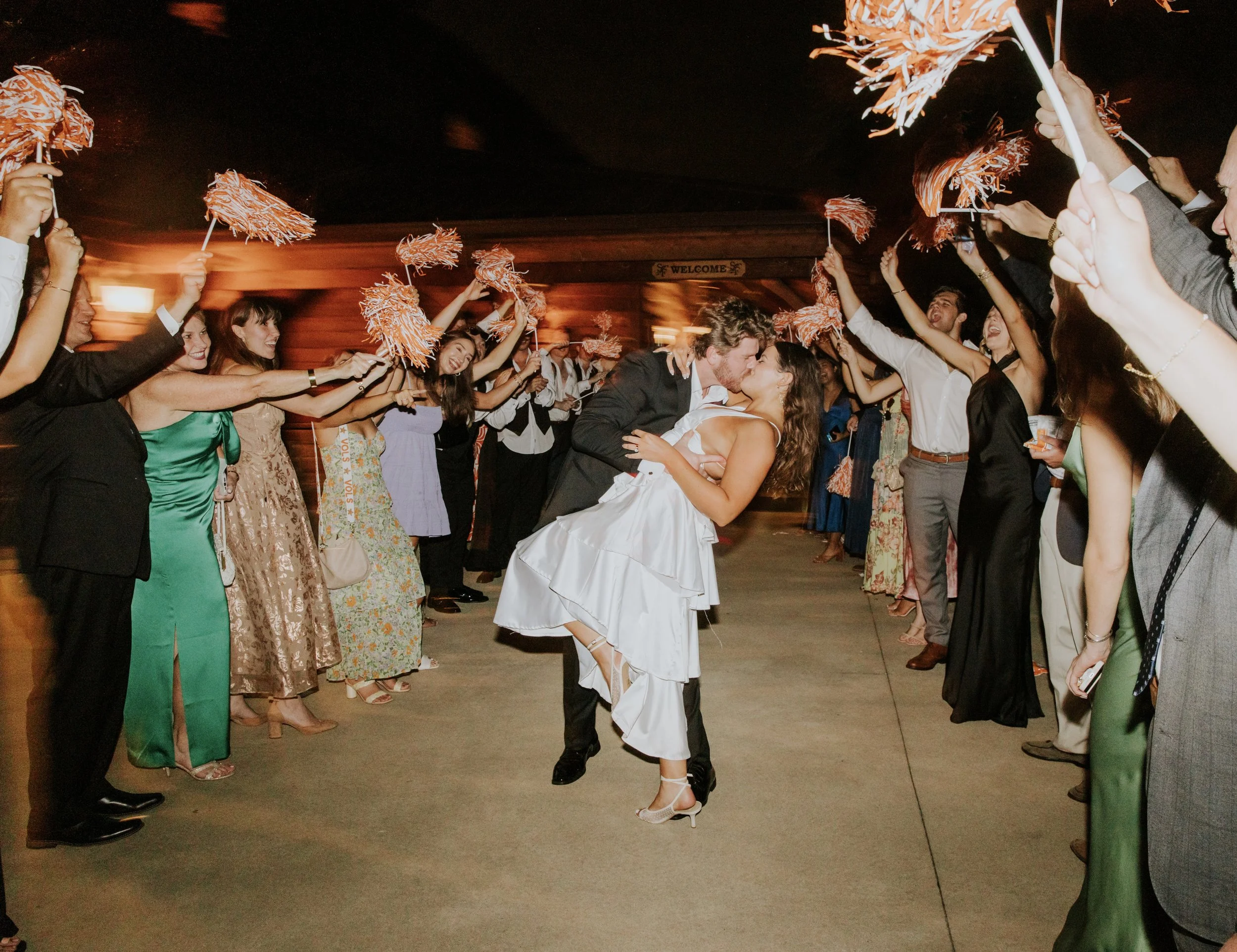 Couple dressed in wedding attire sharing a kiss while being surrounded by guests holding sparklers at night.