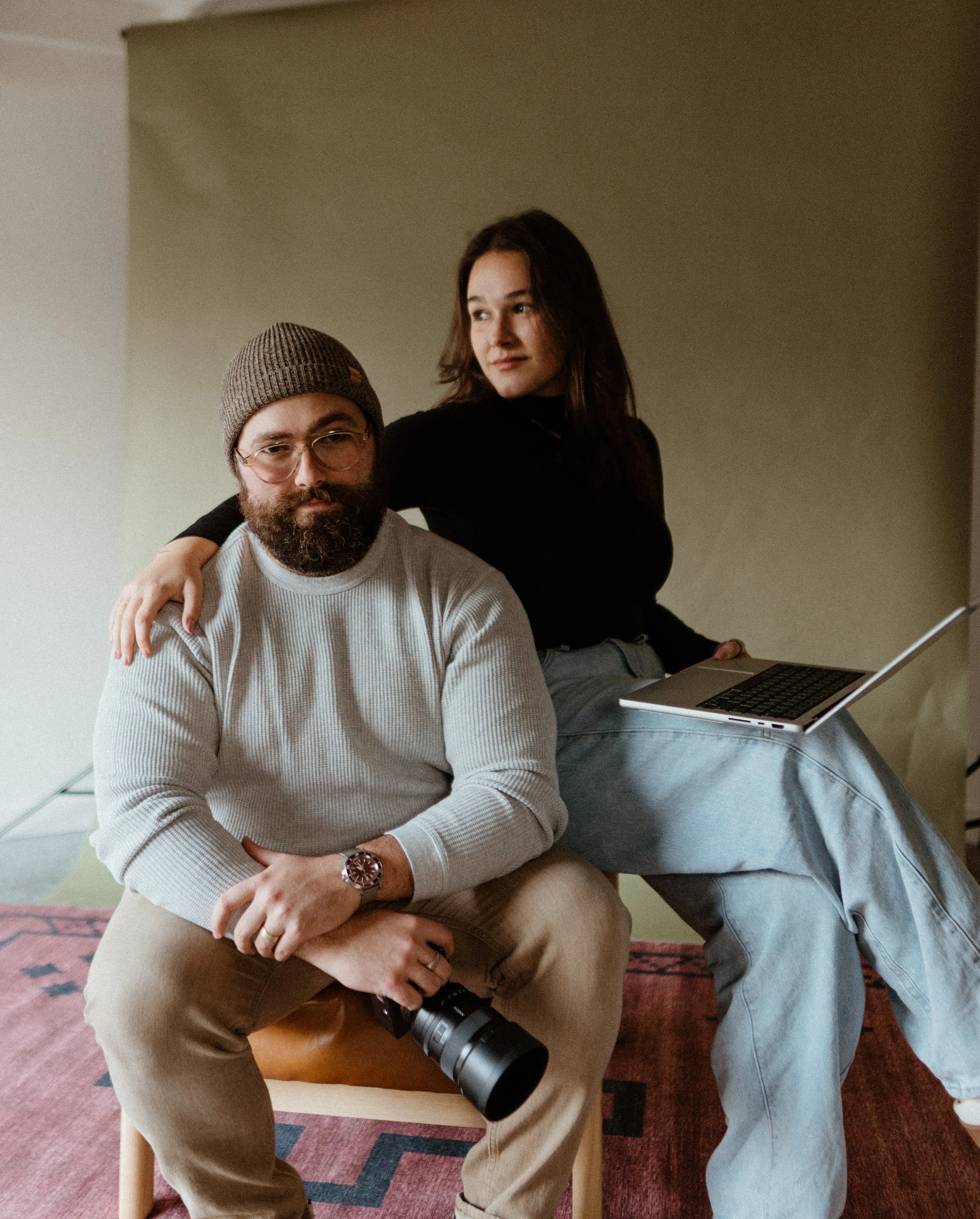 A man with a beard and glasses, wearing a brown beanie and cream sweater, seated on a wooden chair holding a camera. A woman with long dark hair, wearing a black top and gray pants, standing behind him with one arm around his shoulders and holding a laptop on her lap.