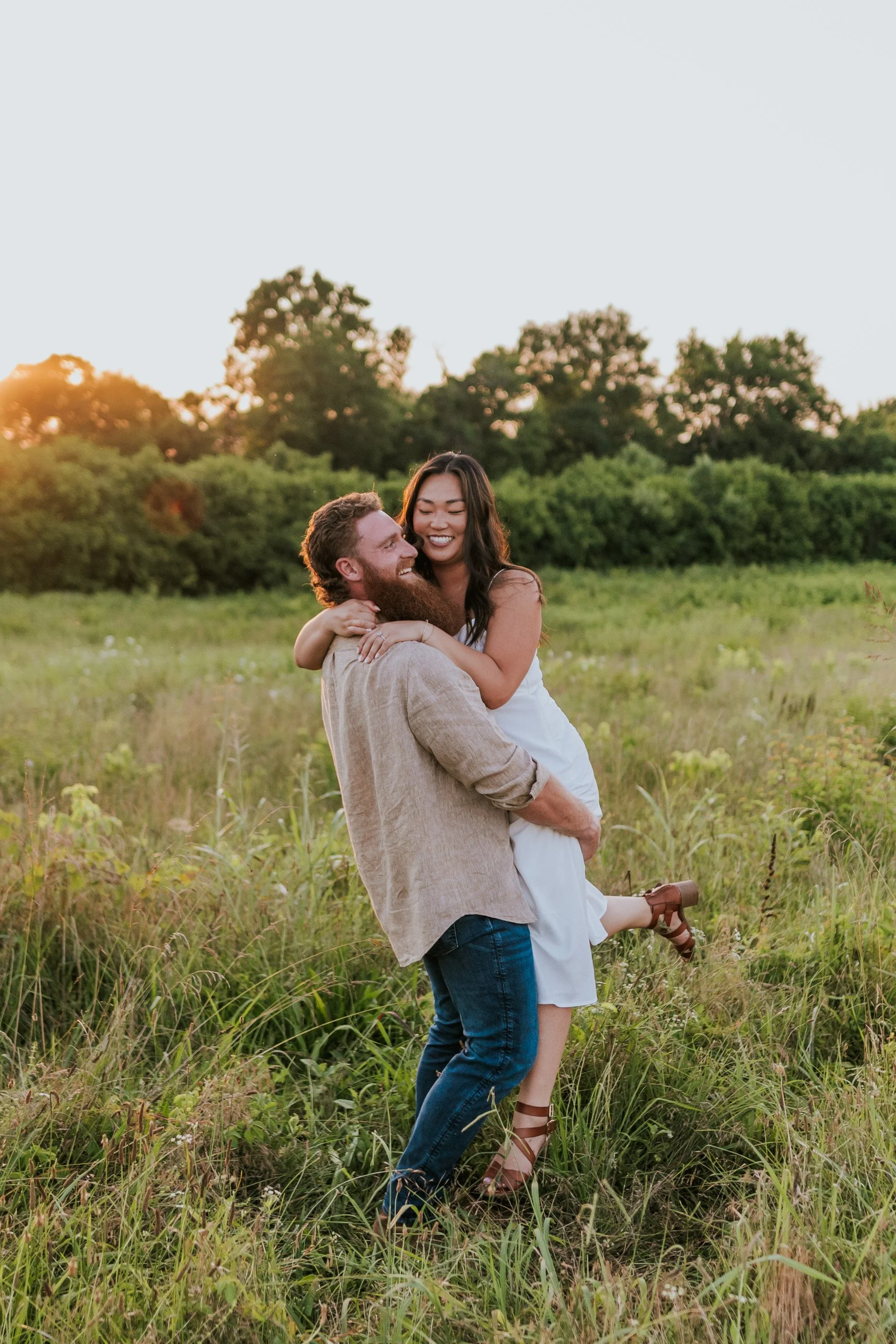 A smiling couple, the man with a beard and the woman with long dark hair, enjoying a playful moment in a grassy field at sunset, with trees in the background.