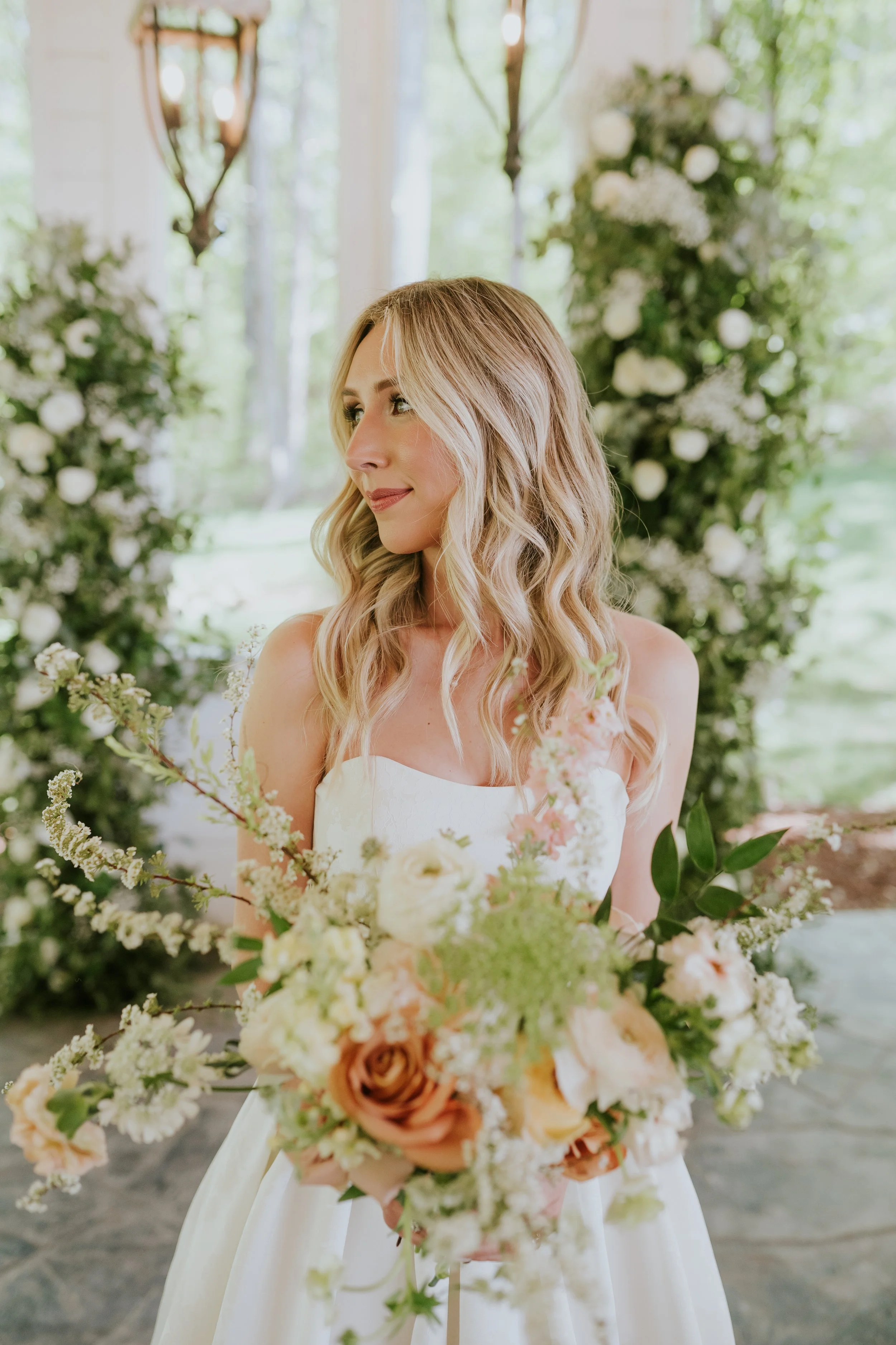 A bride with long, wavy blonde hair in a white wedding dress holding a large bouquet of soft-colored flowers, standing in a decorated outdoor setting with greenery and white flowers.