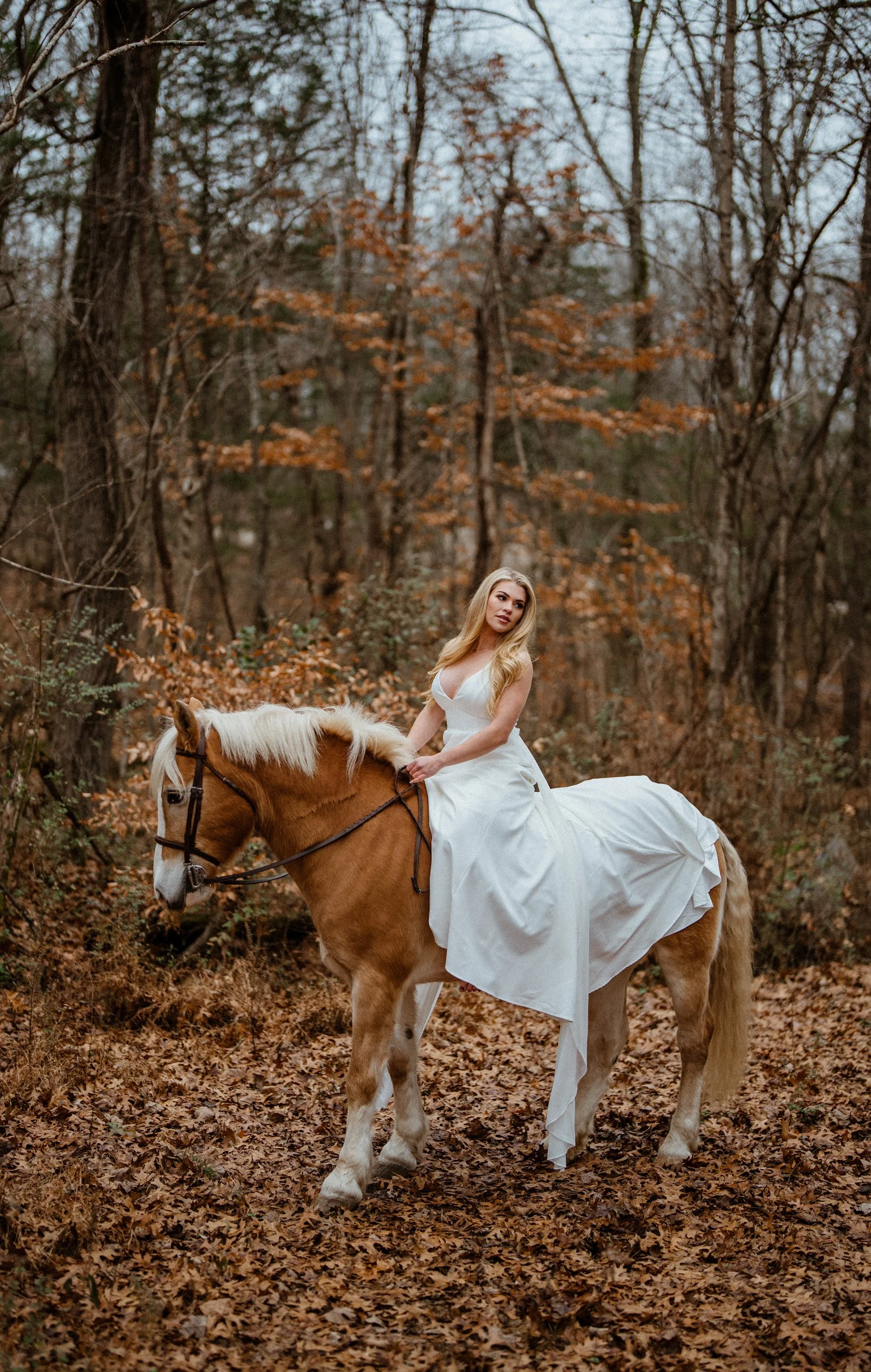 Woman in a white dress riding a light-colored horse through a forest with autumn leaves.