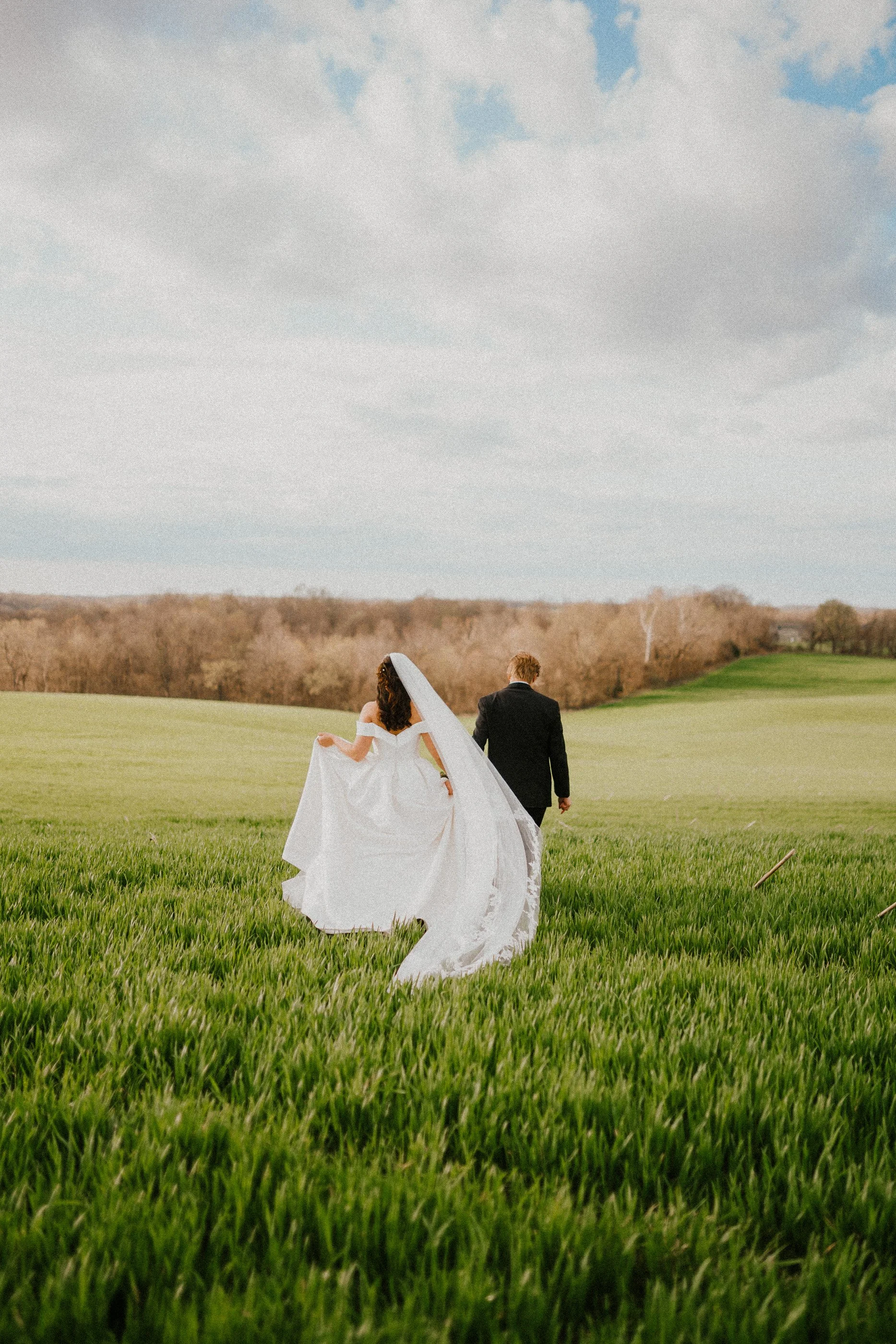 Bride and groom walking away in a grassy field with a cloudy sky.