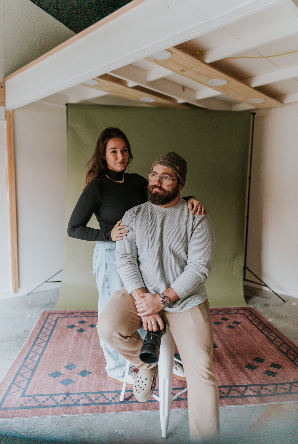 A man with a beard, glasses, and a beanie hat sitting on a white stool, holding a camera lens, with a woman standing behind him, resting her hand on his shoulder in a photo studio with a green backdrop and a red patterned rug.