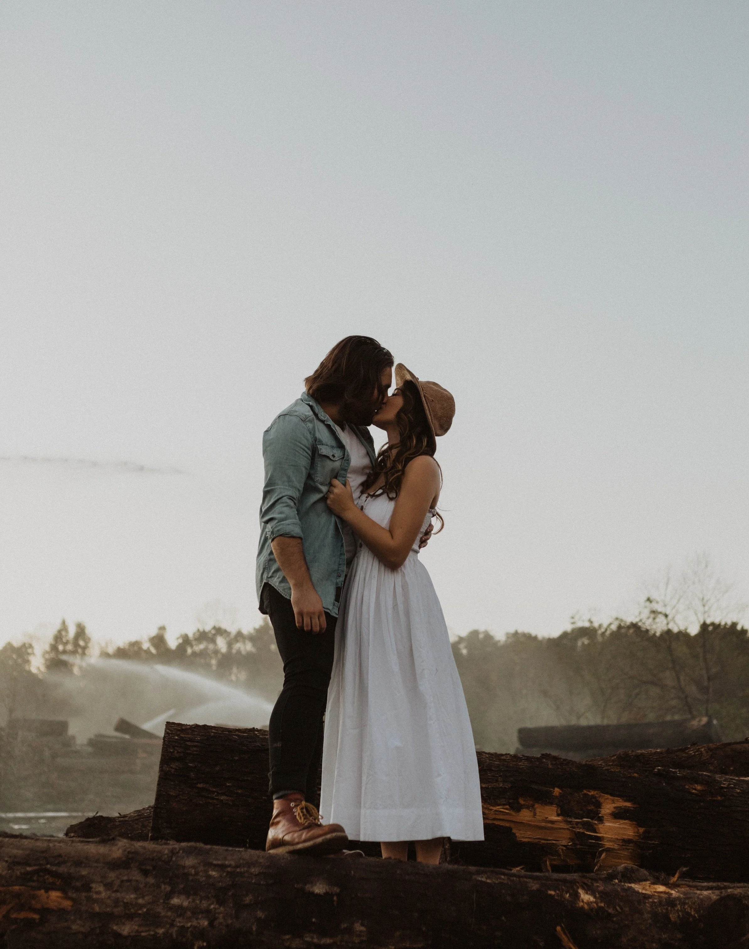 A couple sharing a kiss outdoors on a log with trees and a clear sky in the background.