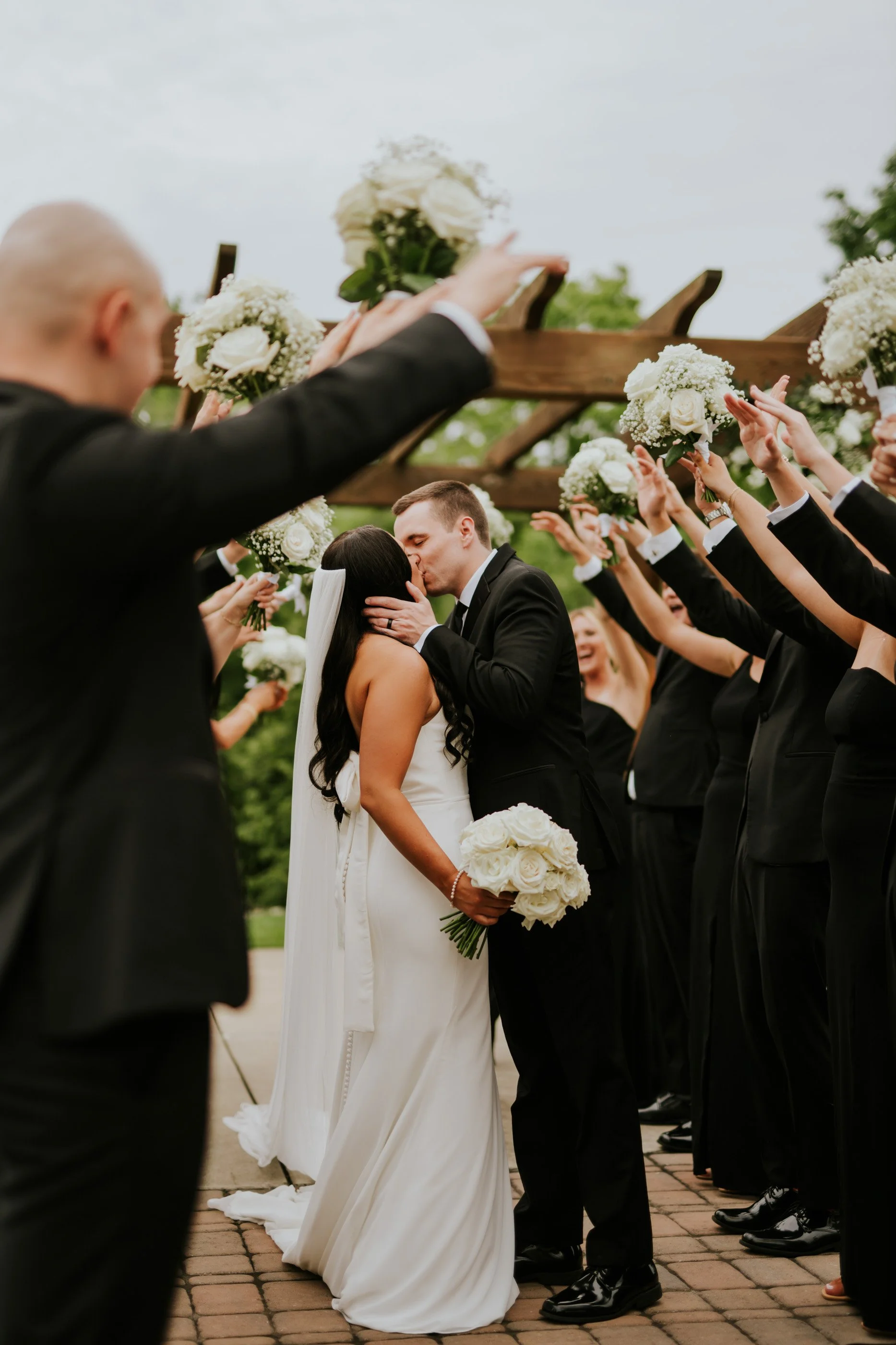 A bride and groom kiss during their wedding ceremony surrounded by bridesmaids and groomsmen holding bouquets and dressed in black tuxedos, outdoors with greenery.
