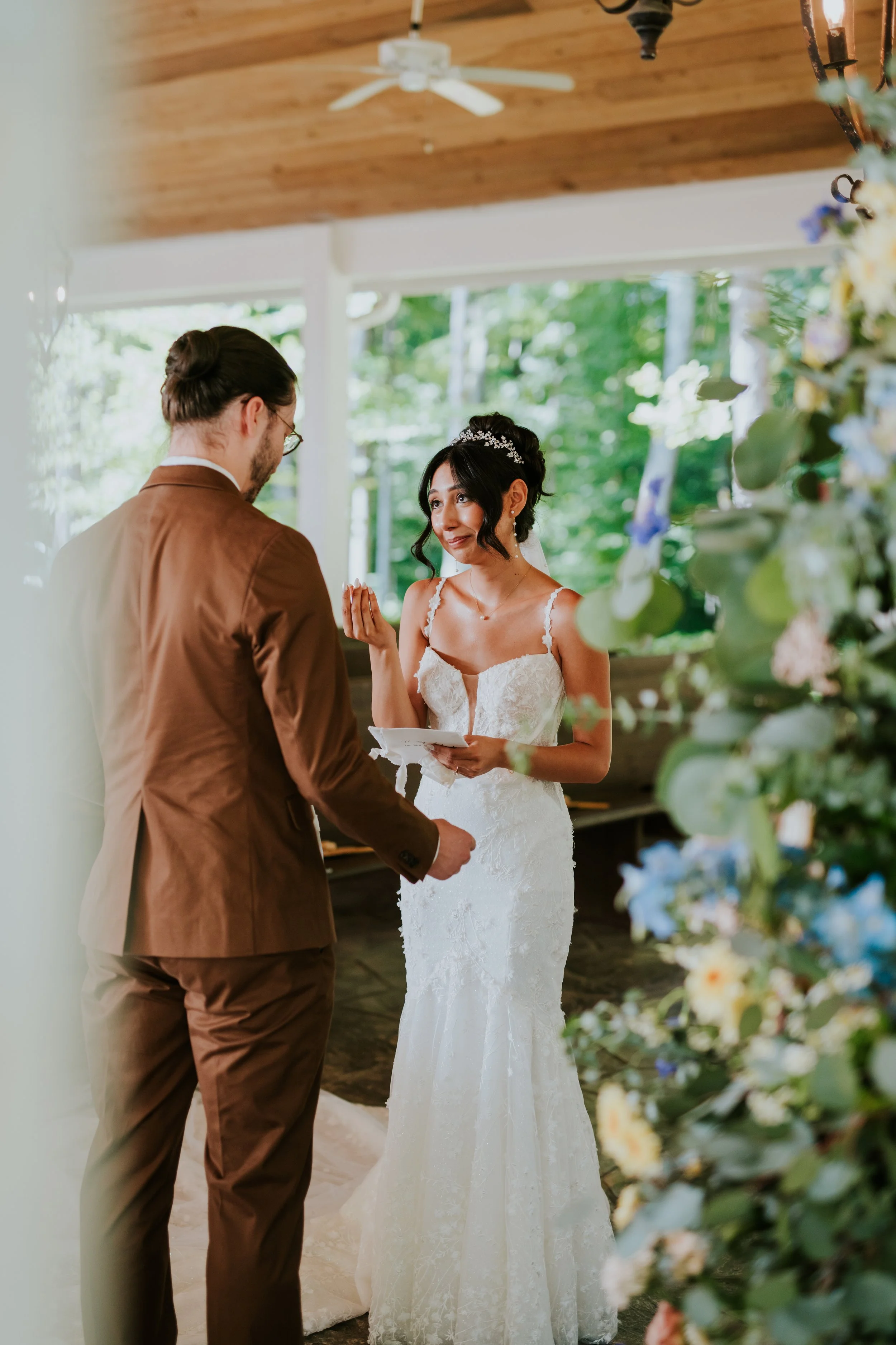 A bride and groom exchanging vows during their wedding ceremony in a bright, green outdoor setting. The bride is dressed in a white lace wedding gown and holding a small plate, while the groom wears a brown suit. The scene includes decorative flowers and a tree canopy in the background.