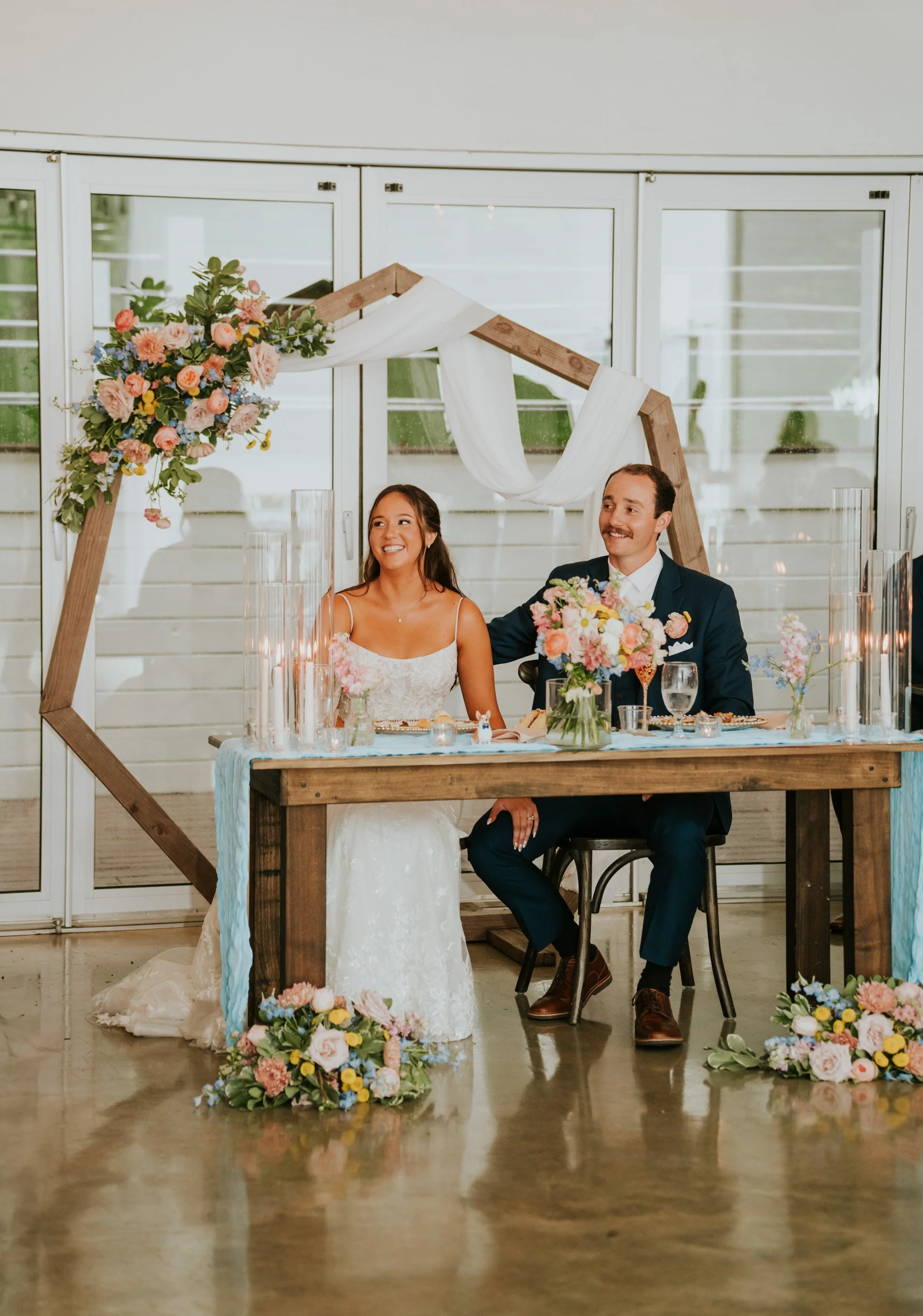 A newlywed couple sits at a decorated reception table with floral arrangements, sitting under wooden decor with draped white fabric, during their wedding reception.