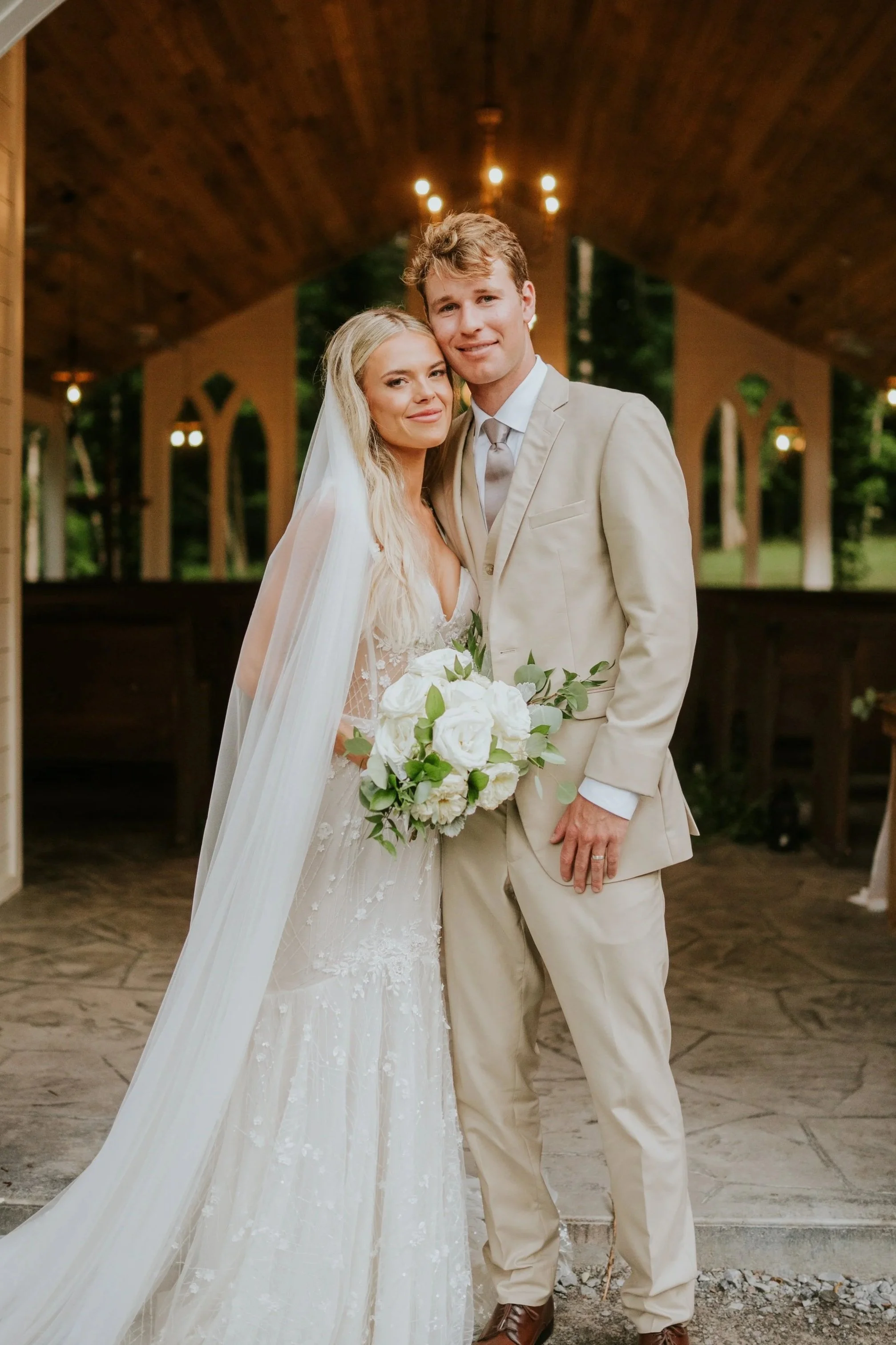 A bride and groom posing together at their wedding, with the bride holding a white bouquet and wearing a white lace wedding dress and veil, and the groom in a light-colored suit. They are standing outdoors under a wooden pavilion with string lights.