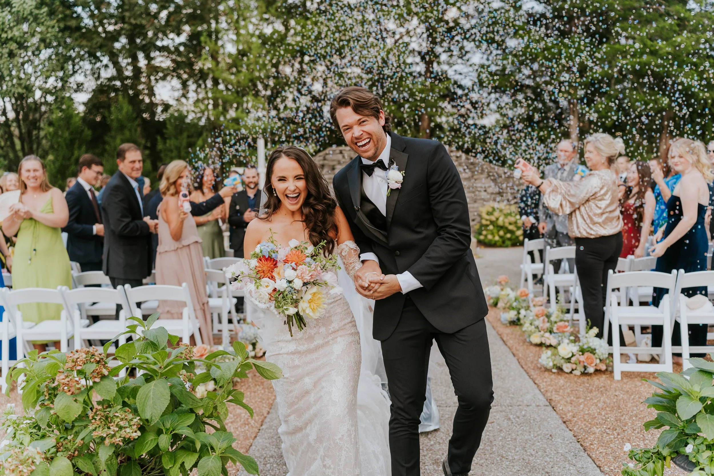 Happy bride and groom at outdoor wedding ceremony, walking down aisle, surrounded by guests, with confetti falling, greenery and flowers in background.