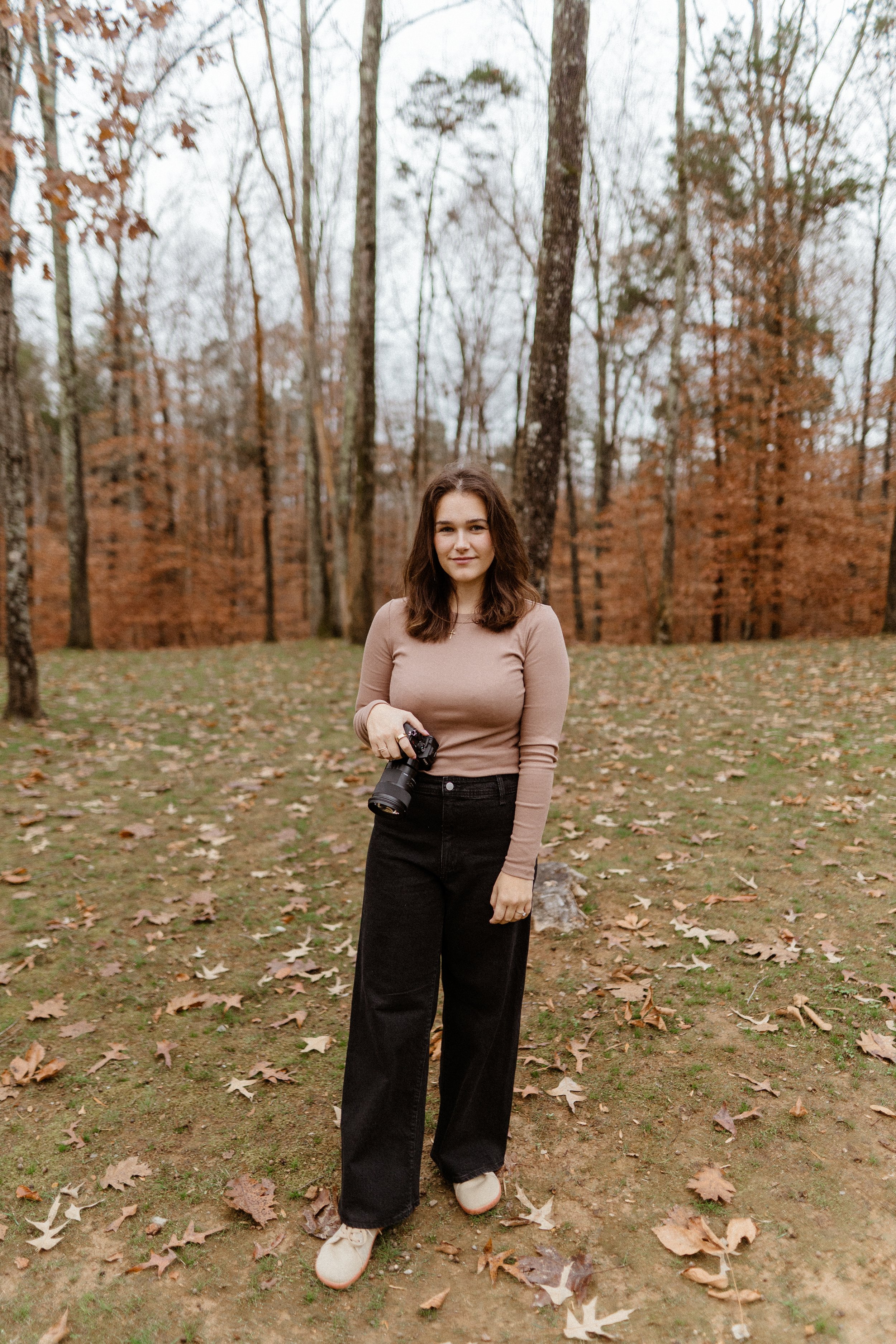 A young woman holding a camera in a forest during autumn, with fallen leaves on the ground and trees with sparse leaves in the background.