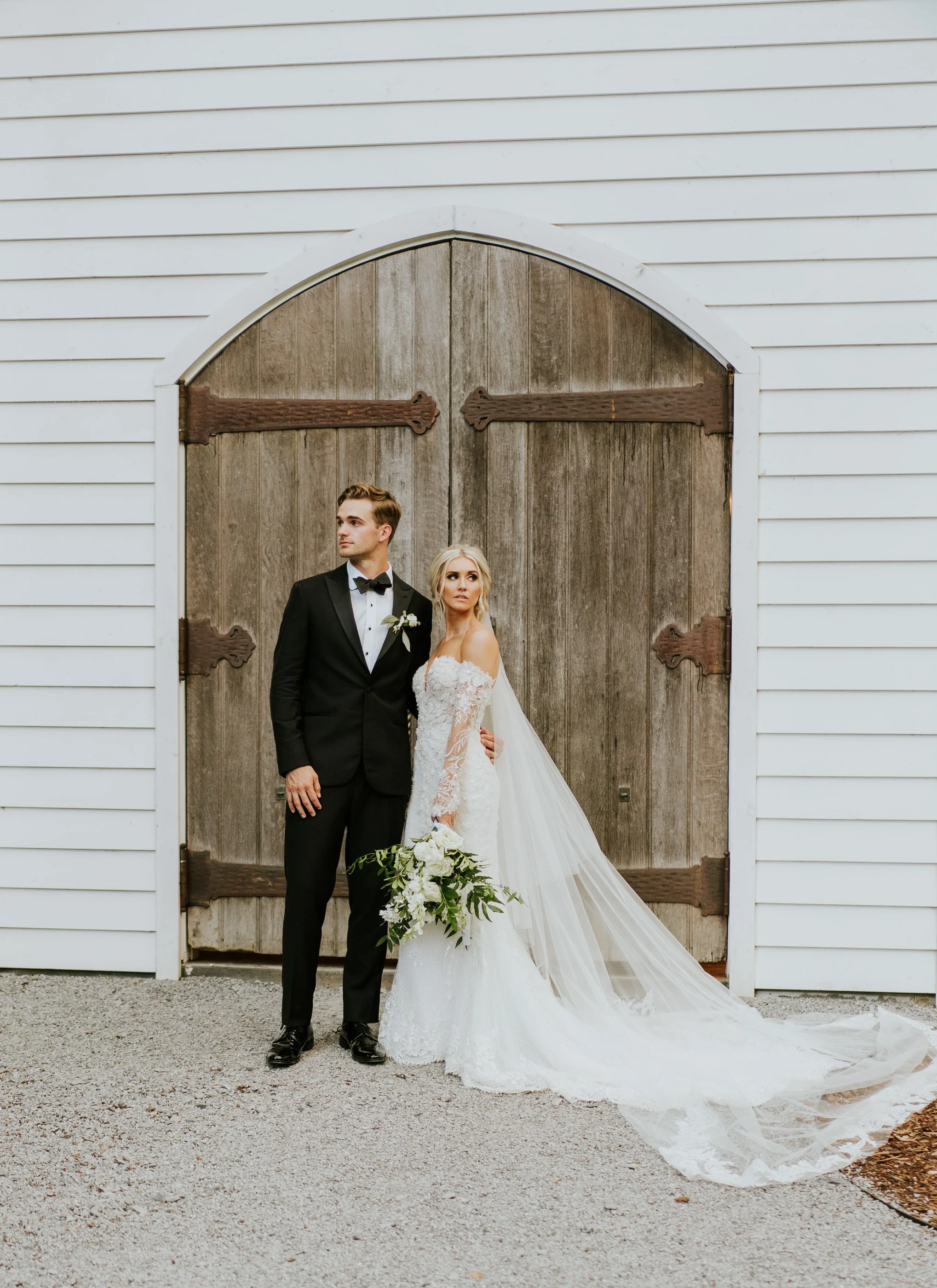 A bride and groom standing in front of a rustic wooden barn door, dressed in wedding attire. The groom wears a black tuxedo with a bow tie, and the bride wears a white lace wedding dress with a long train and holds a bouquet of white flowers and greenery.